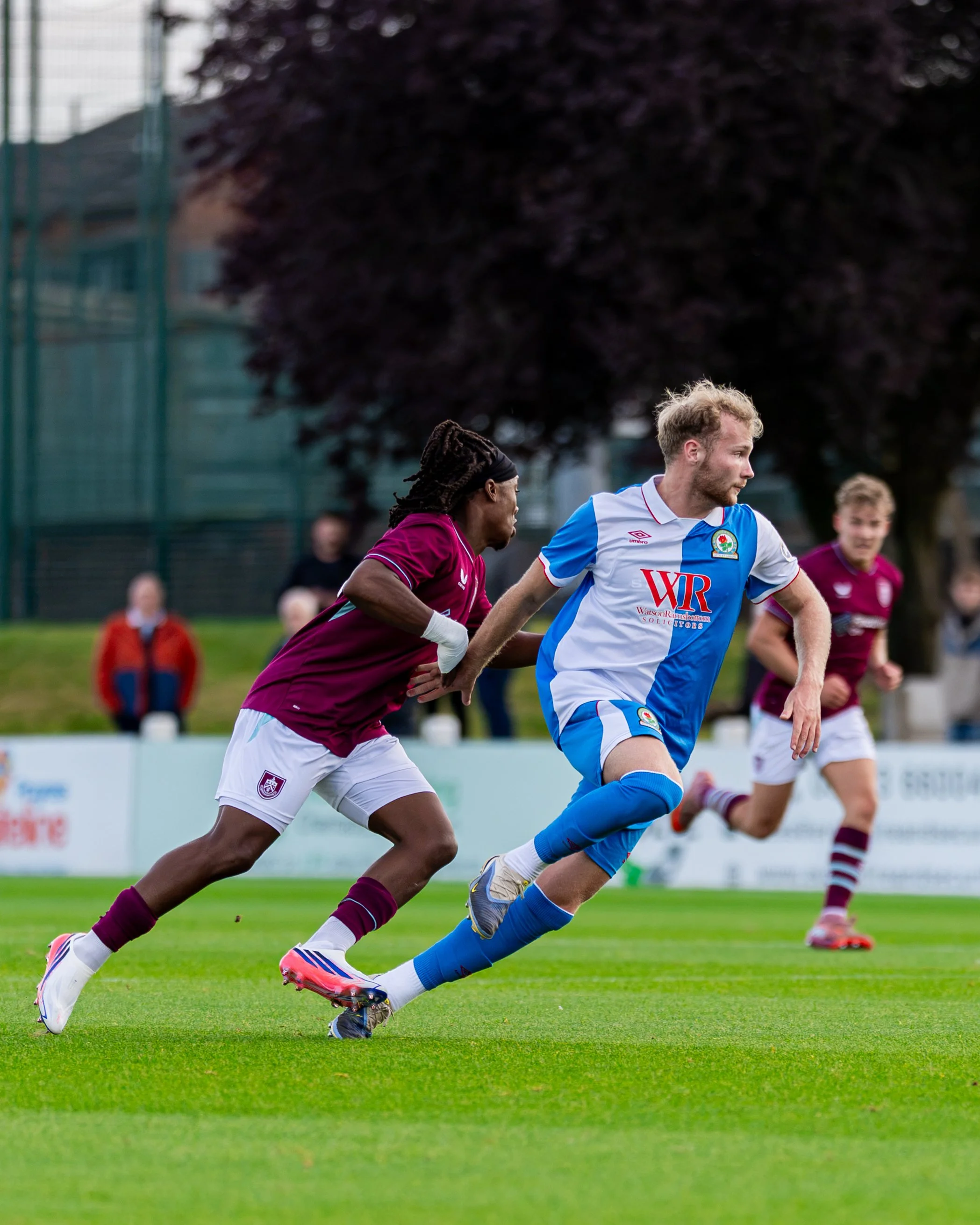 Two soccer players running on a field, one in a maroon uniform and one in a blue and white uniform, with people and trees in the background.