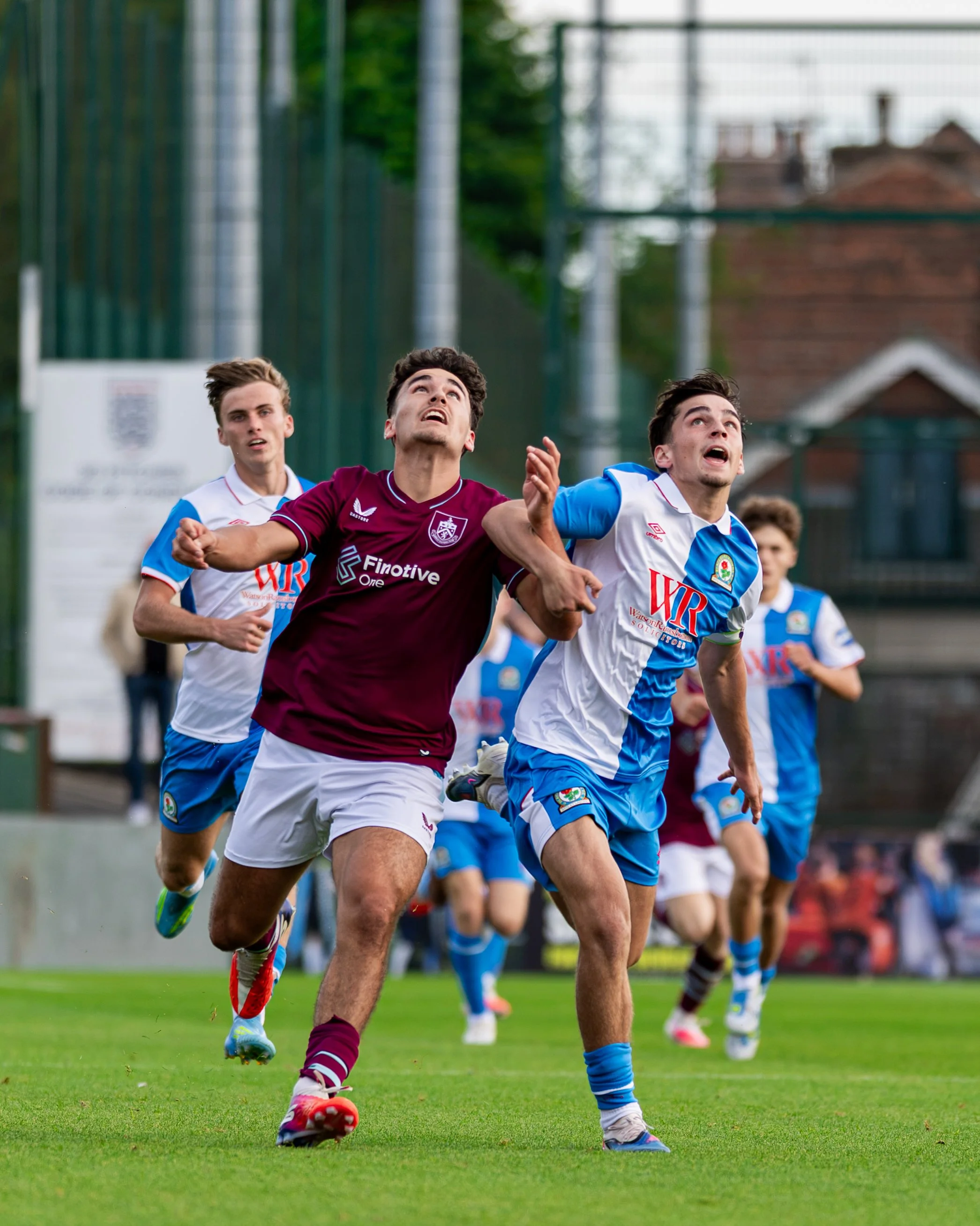 A group of young male soccer players running on a soccer field. One player in a maroon jersey is in front, looking upwards. Others behind him are in white and blue jerseys, also actively moving on the grass field.