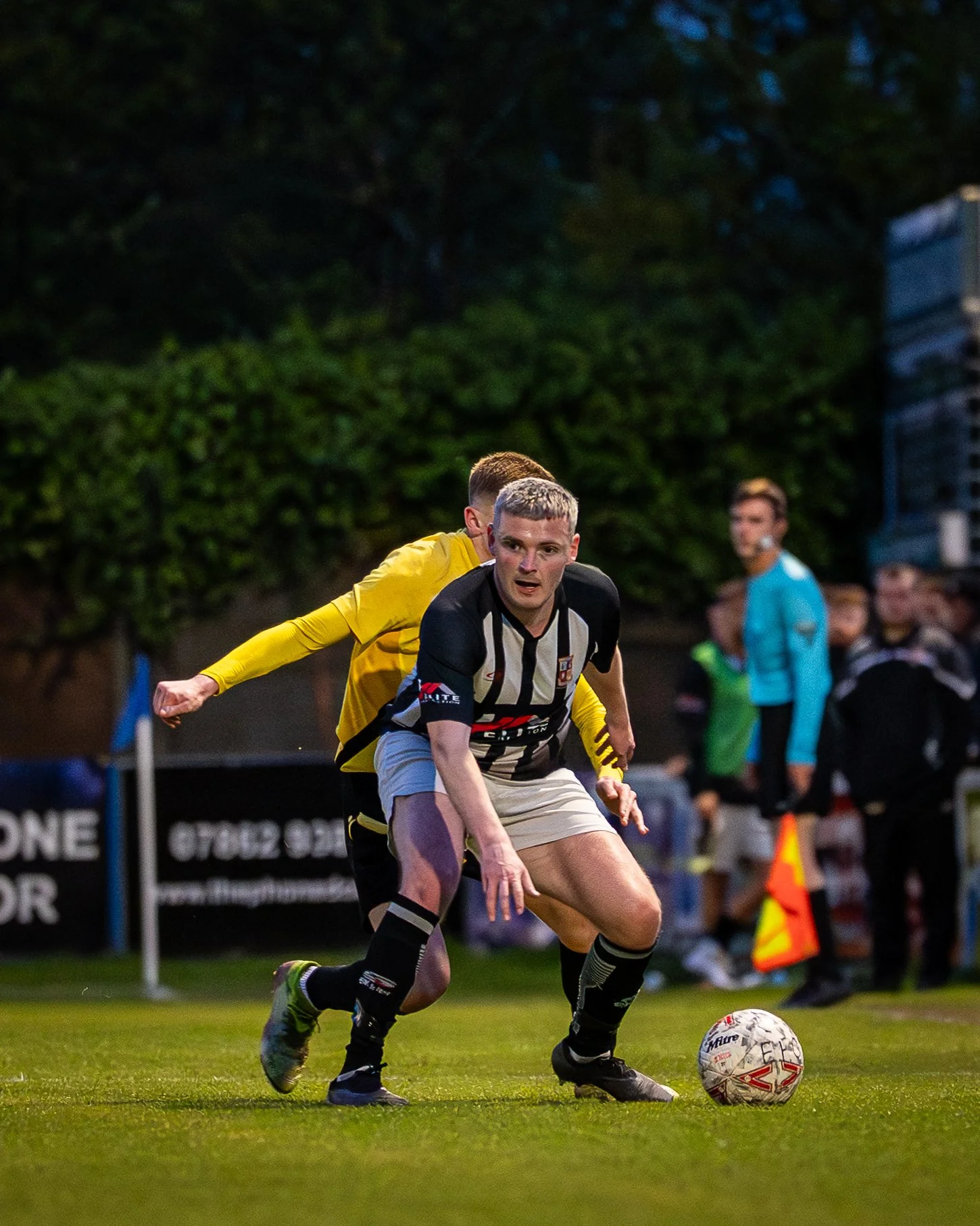 A soccer player wearing a black and white striped jersey and beige shorts is kicking a soccer ball on a field during a game, with another player in a yellow jersey close behind and referees and spectators in the background.