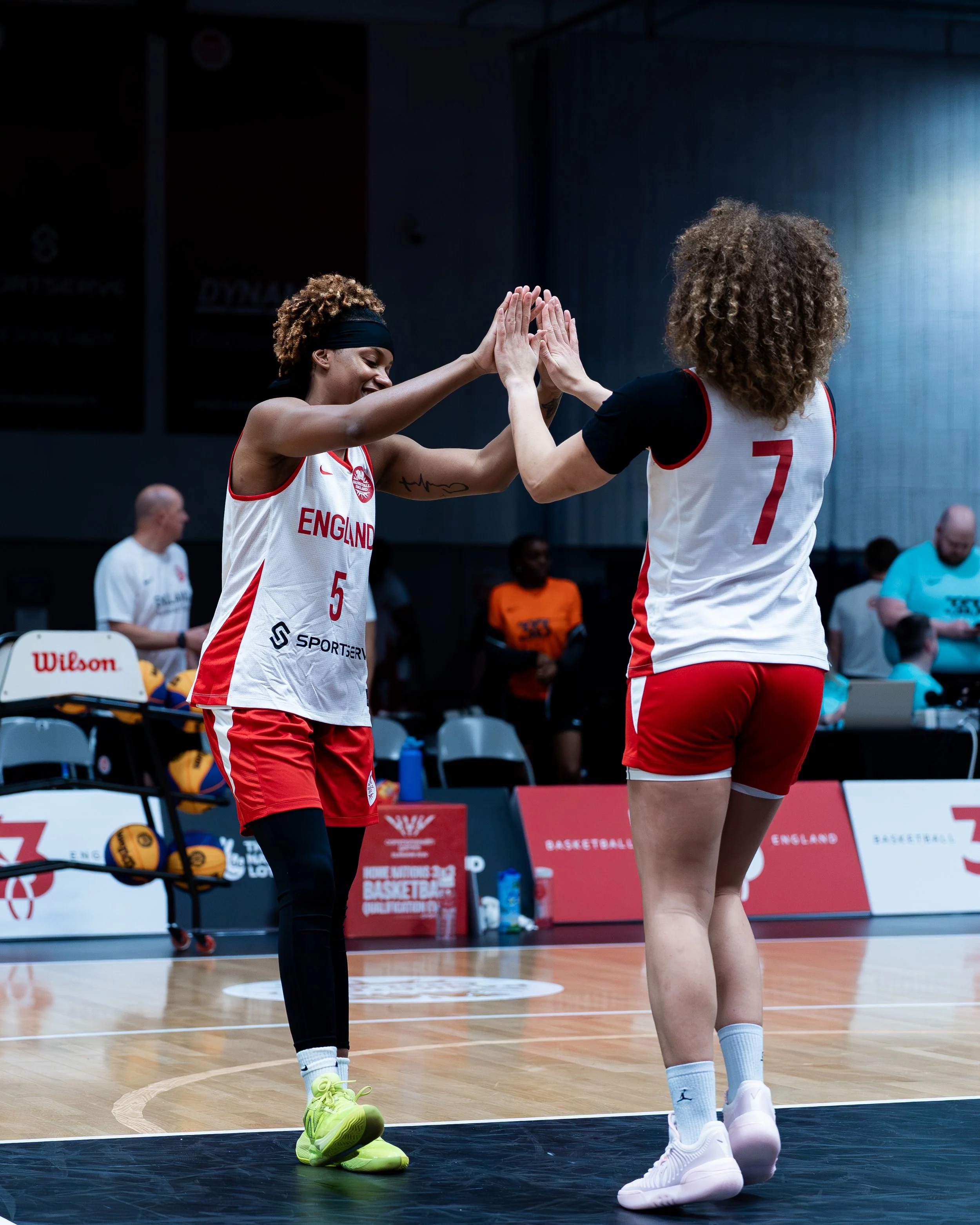 Two female basketball players from England high five during a game on a basketball court. One wears a jersey with the number 5 and the other with the number 7. Both have curly hair and are dressed in athletic gear with sneakers.