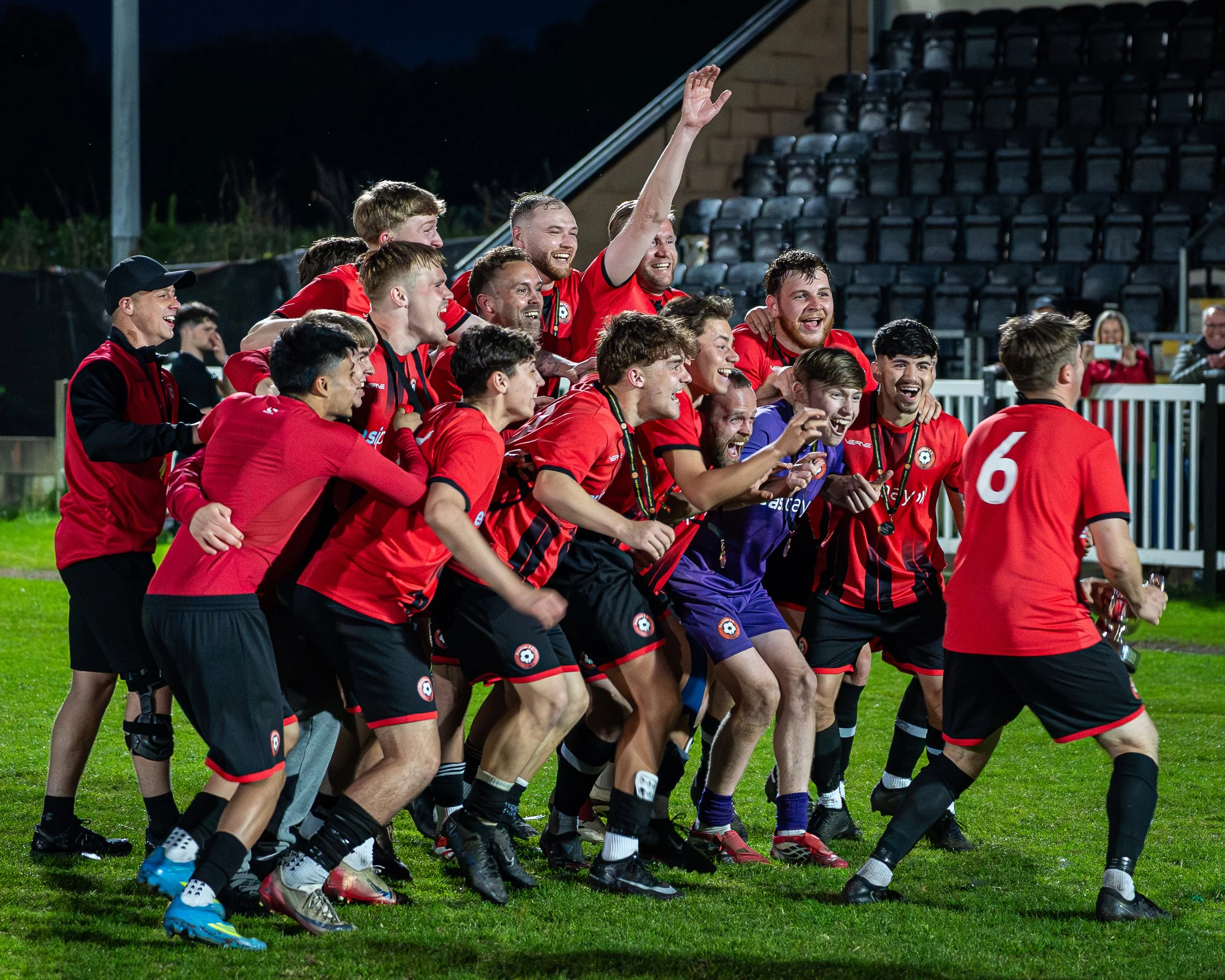 A group of soccer players in red uniforms celebrating on the field after a game, with some players smiling and hugging, and a woman taking a photo in the background.