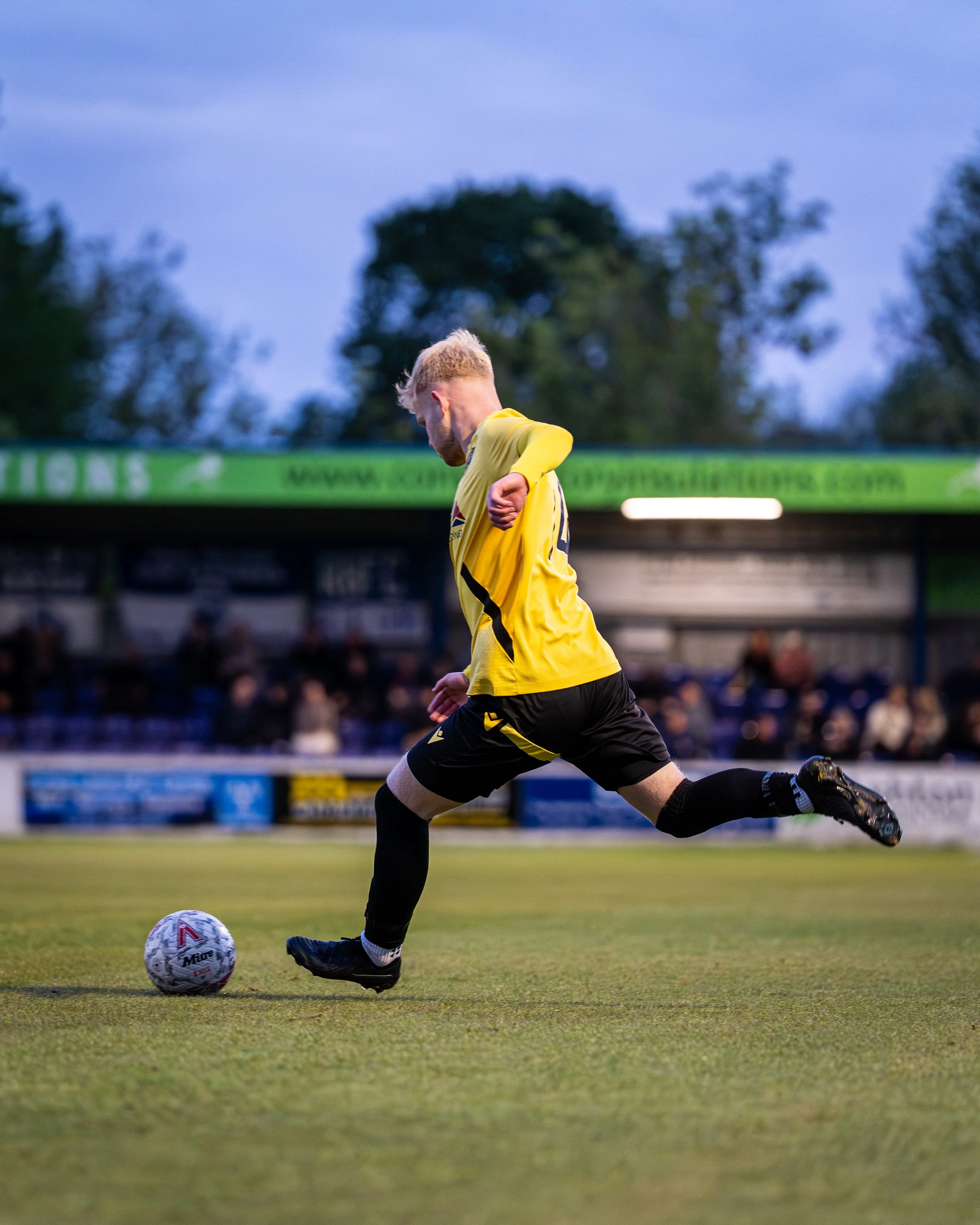 A soccer player in a yellow jersey and black shorts is kicking a soccer ball on a grass field during a game, with trees and a blurred crowd in the background.
