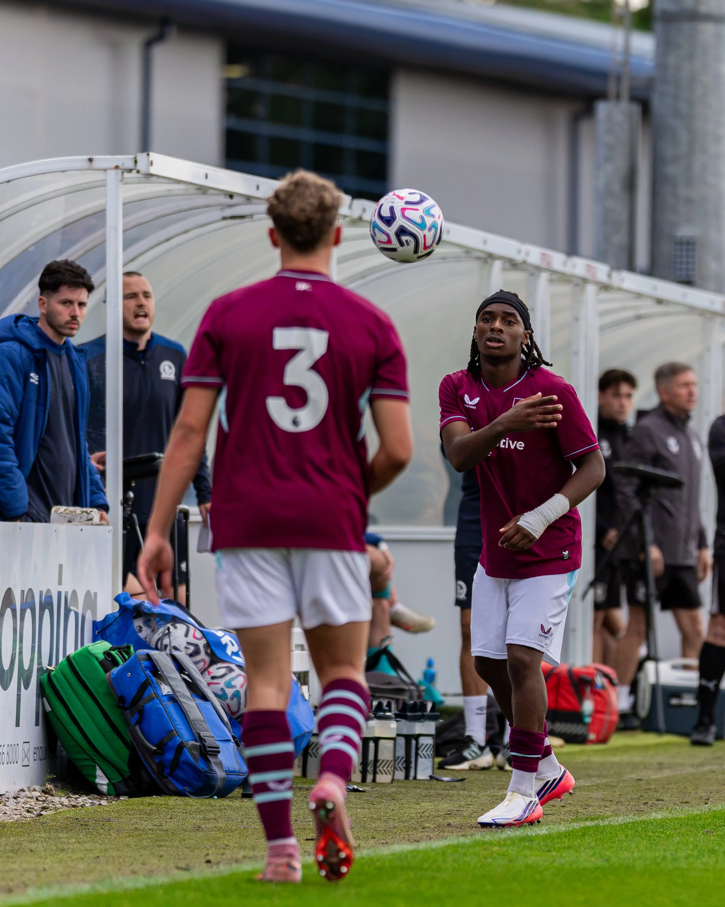 A soccer player in a maroon jersey with the number 3 is warming up or practicing on the sidelines of a soccer field, with other players and staff members standing nearby.