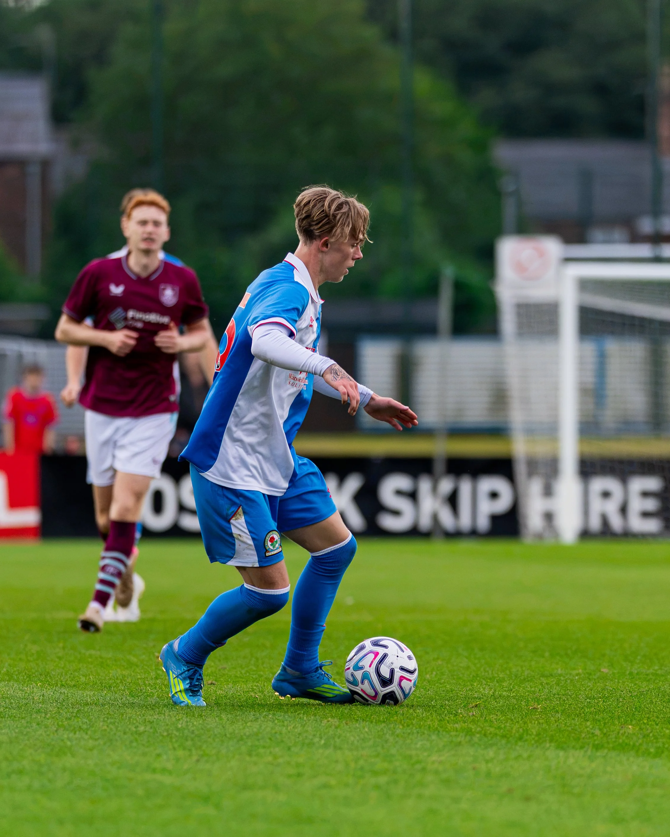 A soccer player in a blue and white jersey is about to kick a soccer ball on a grass field. Another player in a maroon jersey is running in the background.