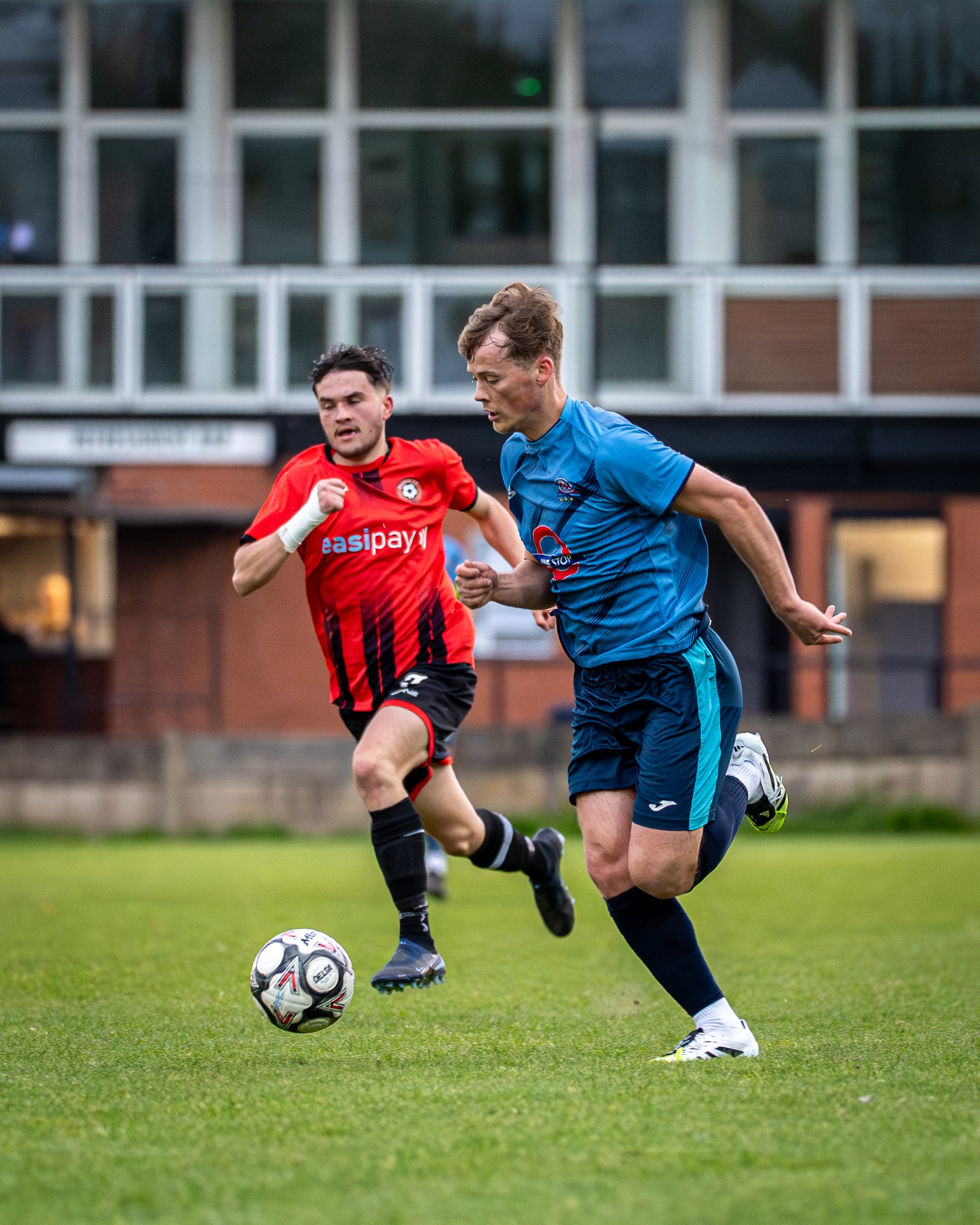 Two soccer players are competing for the ball on a grassy field, one in a red jersey and the other in a blue jersey, with a building in the background.