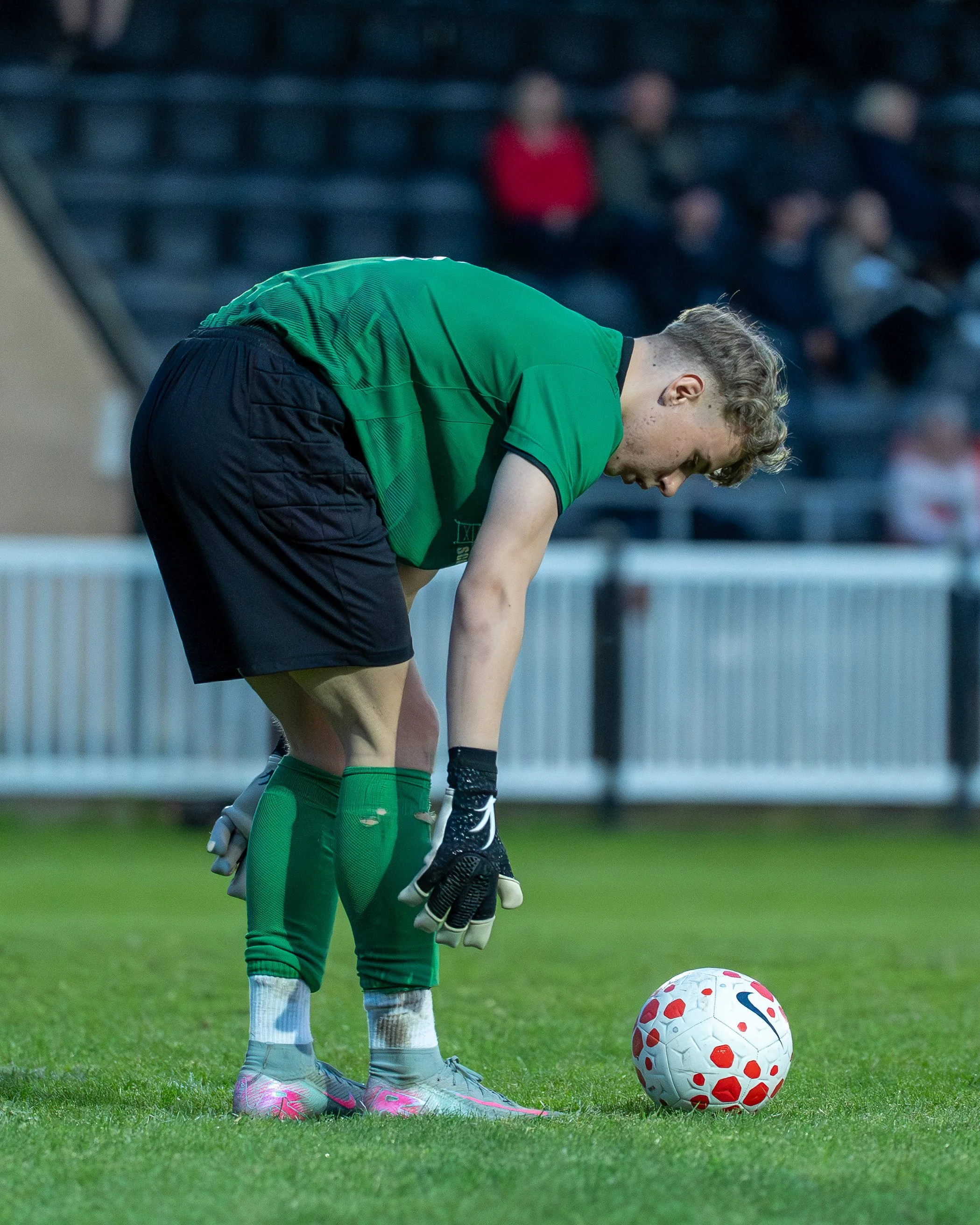 A young soccer player in a green jersey and gray shorts is bent over on the field, preparing to kick a white and red soccer ball. The player is wearing pink and gray cleats and green socks. In the background, spectators are sitting in the stands, som