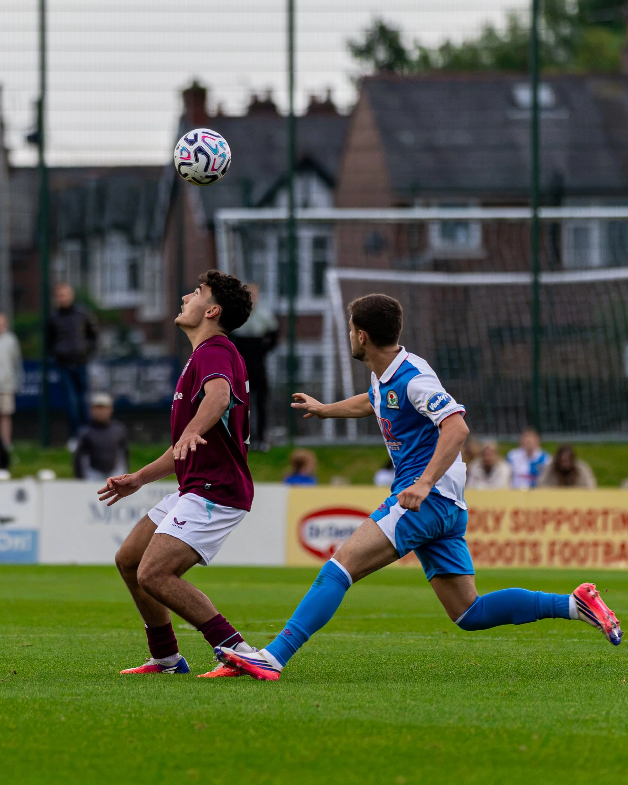Two soccer players competing for the ball during a game on a green field with residential houses in the background. One player is wearing a maroon jersey and white shorts, the other is in a blue jersey and matching shorts.