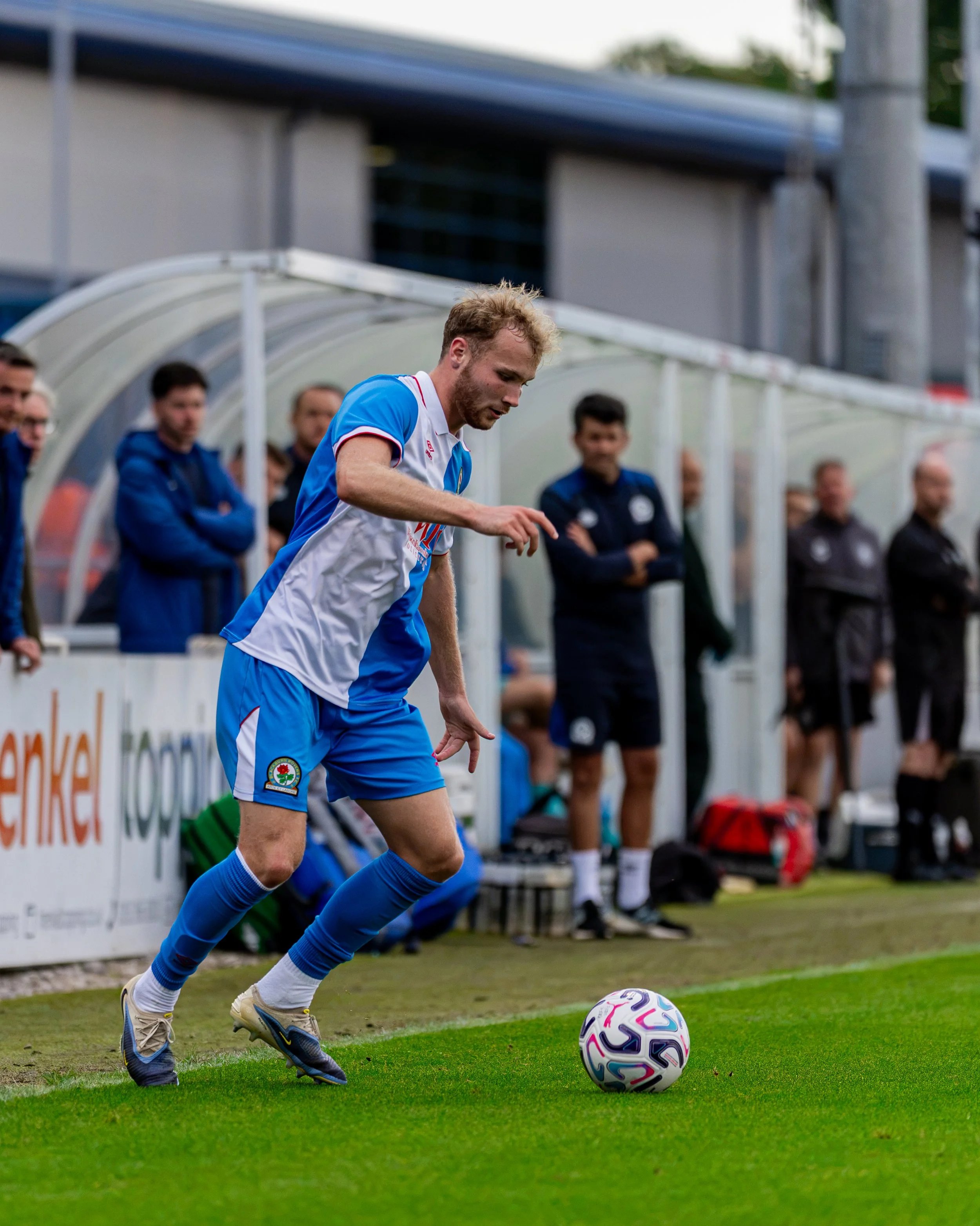 A male soccer player in a blue and white uniform is kicking a soccer ball on the field, with a dugout and several spectators in the background.