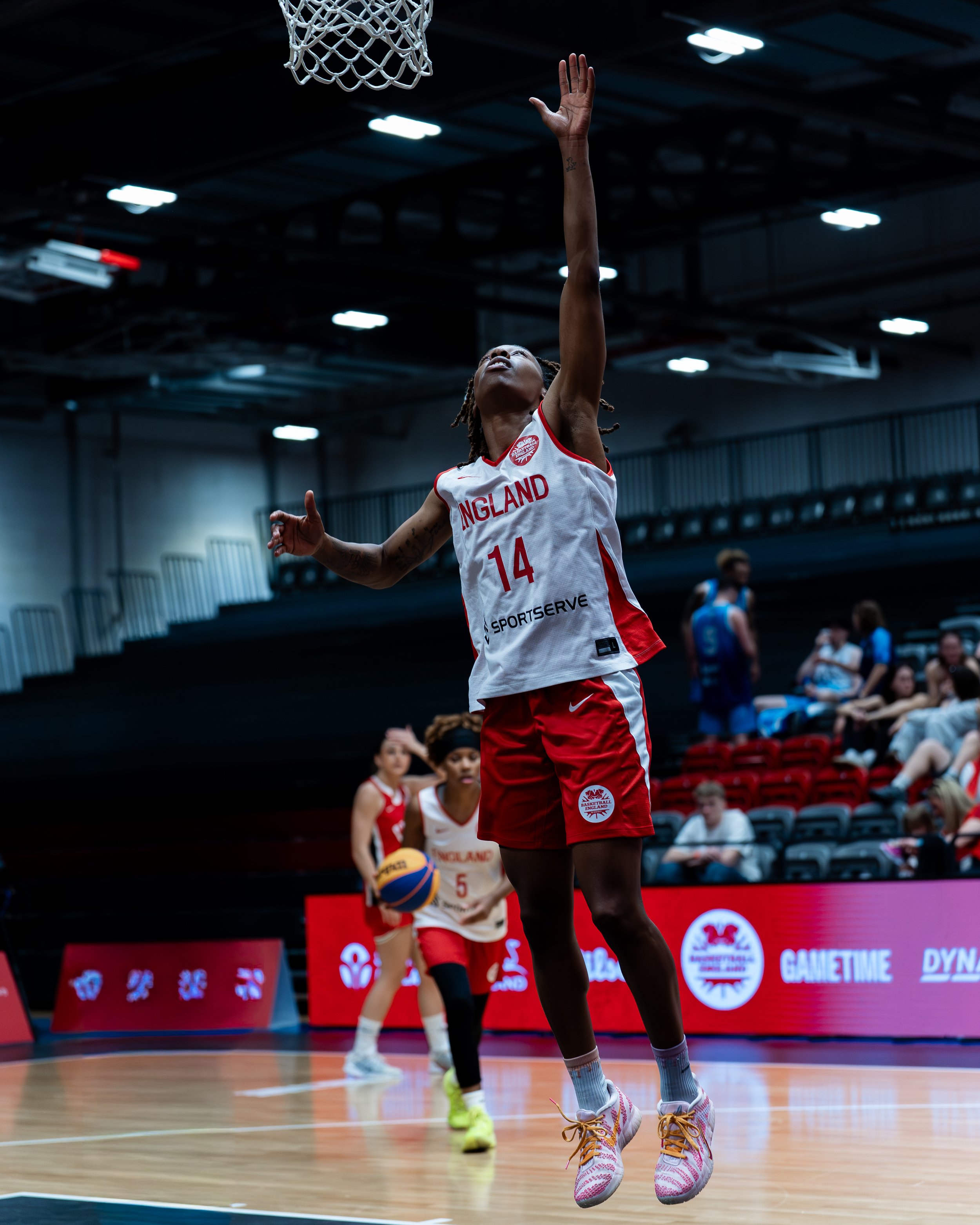 An England women's basketball player wearing jersey number 14 jumps towards the basketball hoop during a game, with other players and spectators visible in the background.