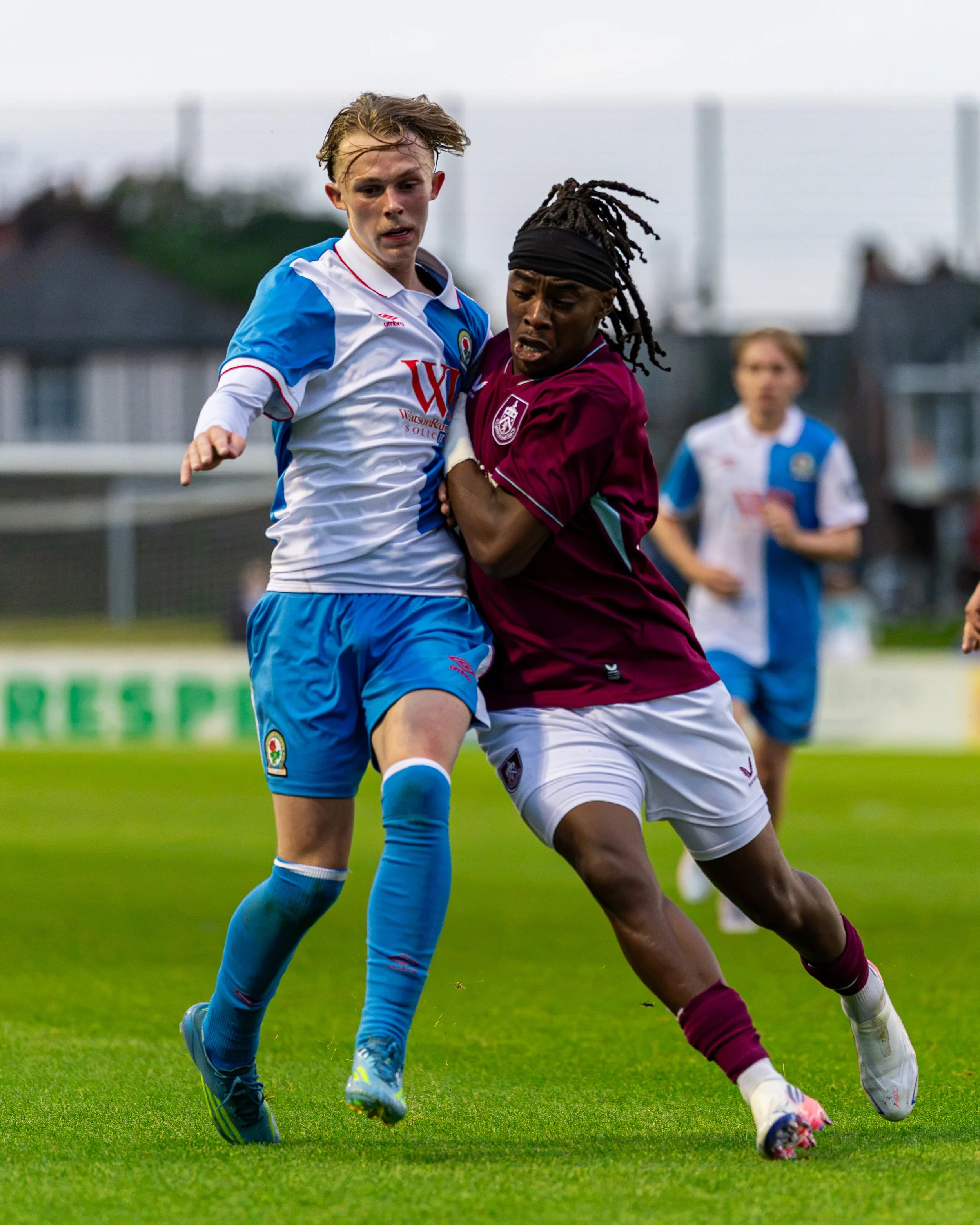 Two soccer players competing for the ball on a green field, one wearing a white and blue jersey and the other in a maroon jersey, with a blurred background of other players and houses.