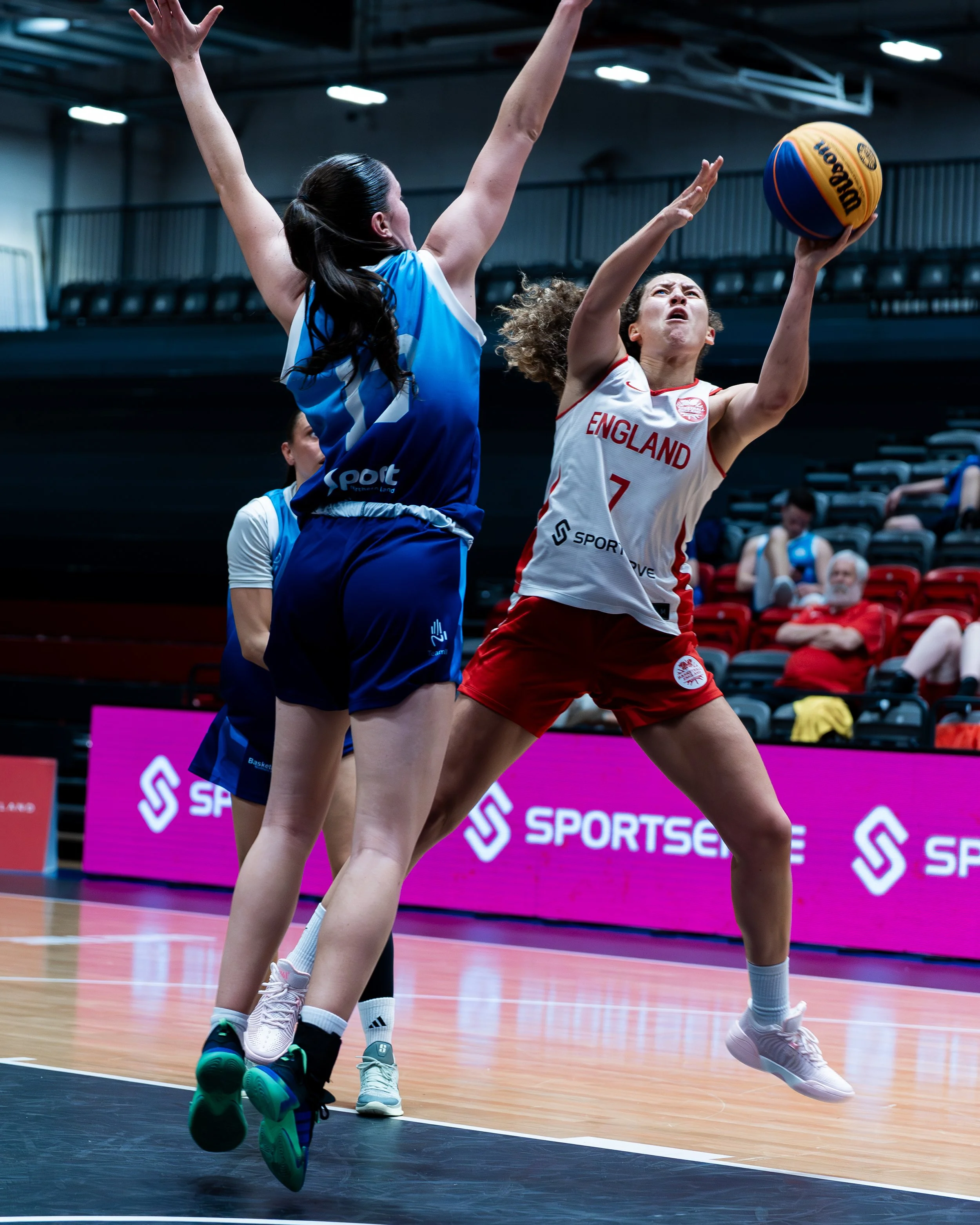 Two female basketball players are jumping for the ball during a game. One player, wearing a white jersey with “England” and the number 7, is reaching for the ball. The other player, in a blue uniform, is defending. The arena has seated spectators and