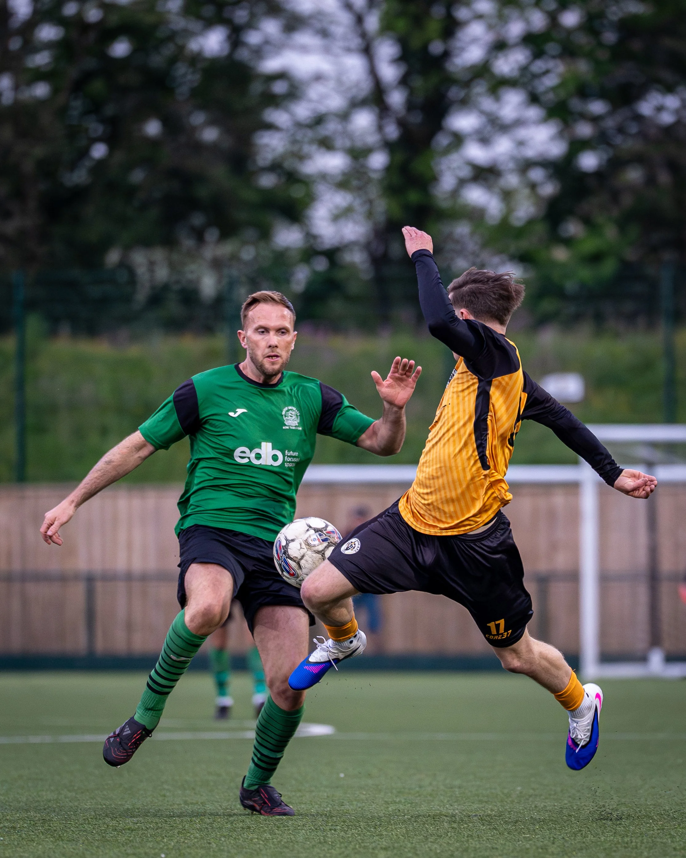 Two soccer players competing for the ball during a match on a green field, with one in a green jersey and the other in a yellow and black jersey, both focused on the ball.