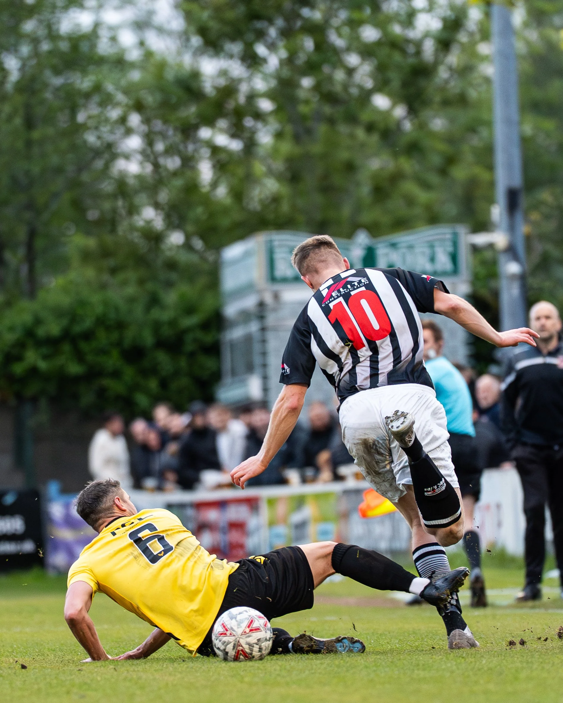 A soccer match scene where a player in a black and white striped uniform is jumping over a goalkeeper in a yellow jersey, who is on the ground extending his leg to block a shot on goal. The background shows a crowd of spectators and greenery.