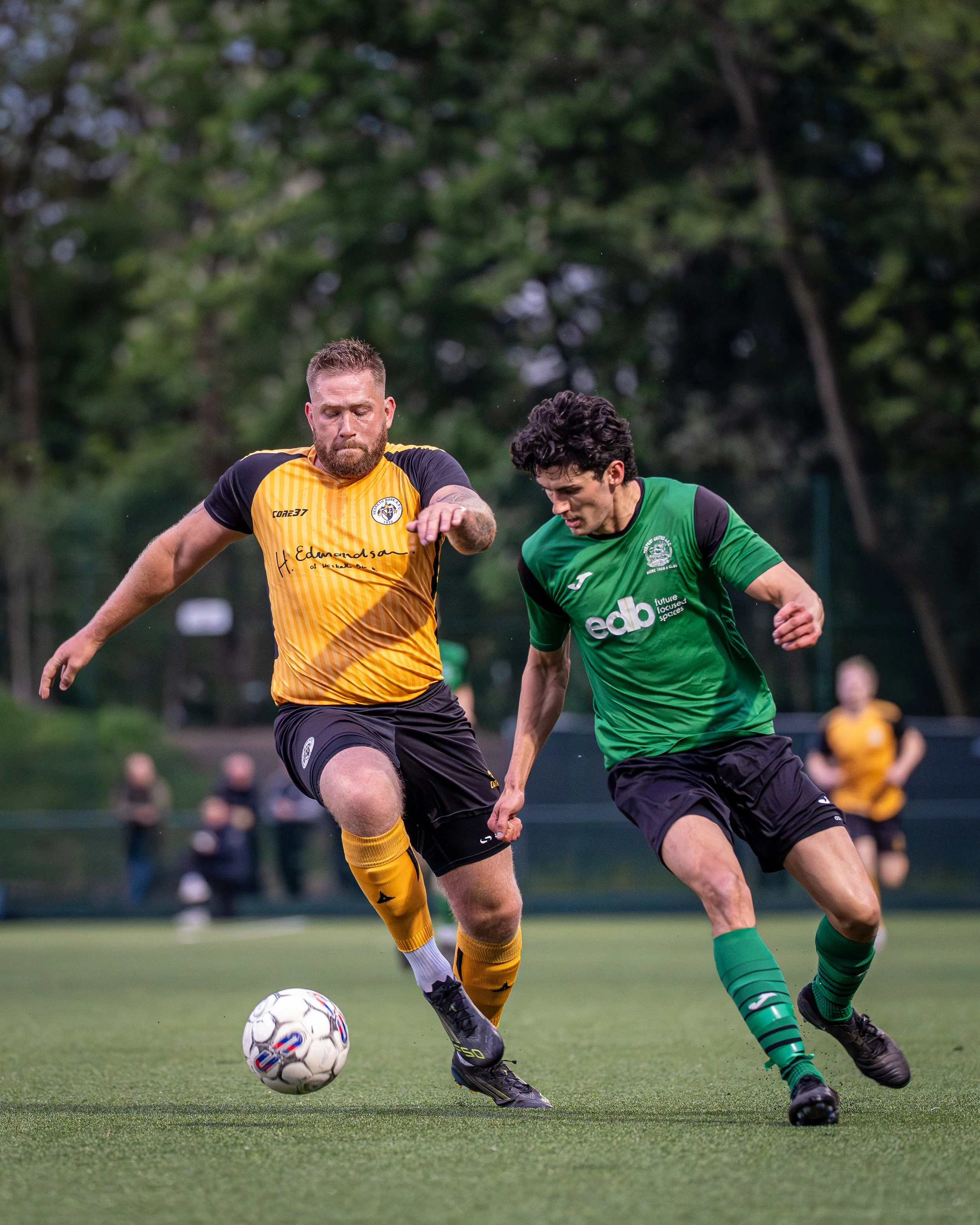 Two soccer players racing for the ball on a green field during a match, with blurred spectators in the background.