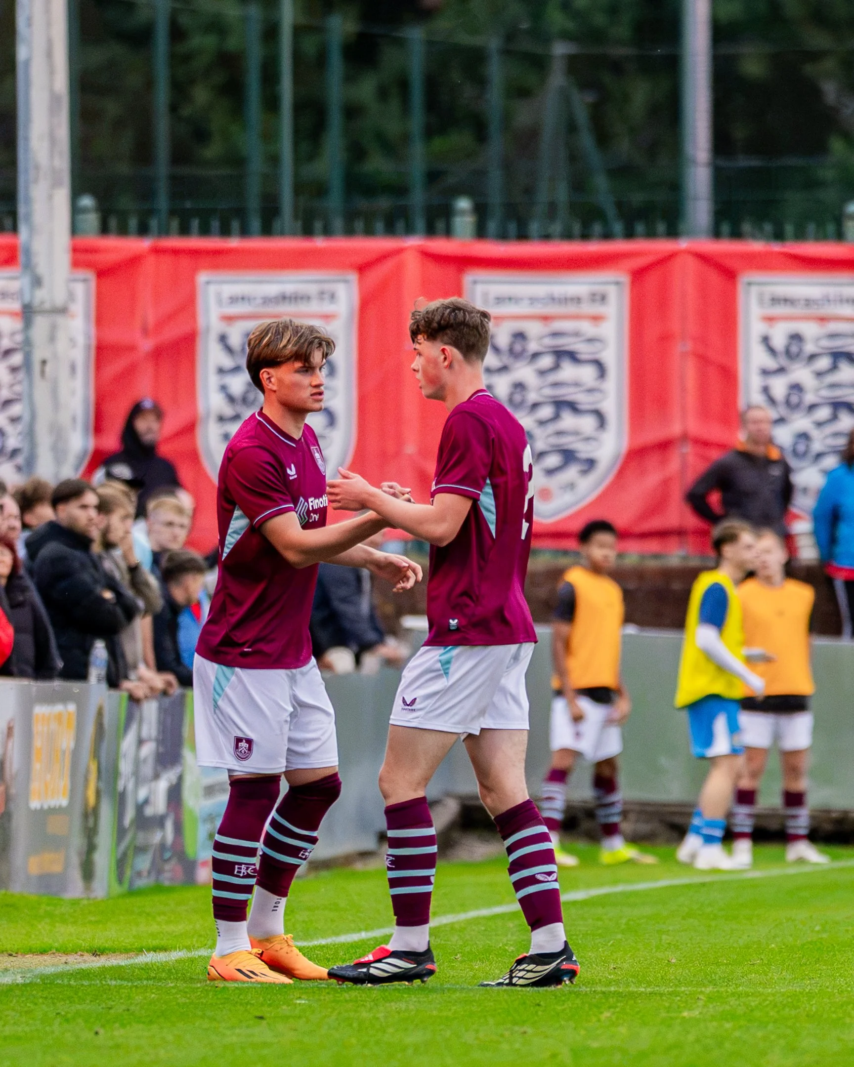 Two soccer players in maroon jerseys and white shorts shaking hands on the field during a game. Spectators and team staff are visible in the background.