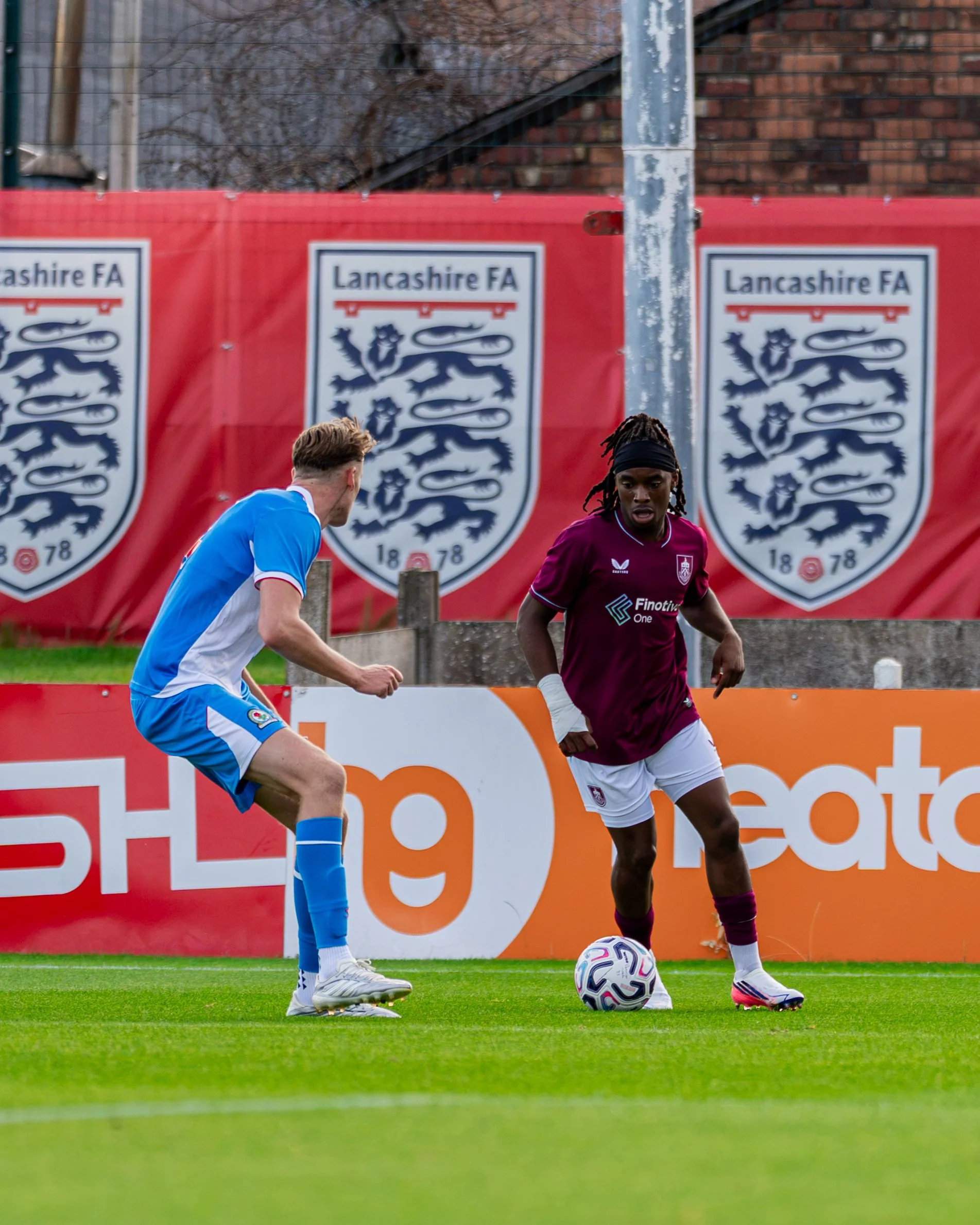 Two soccer players, one in a blue uniform and one in a maroon uniform, are on a soccer field near the sideline, with a soccer ball between them. Behind them are banners with the Lancaster FA logo.