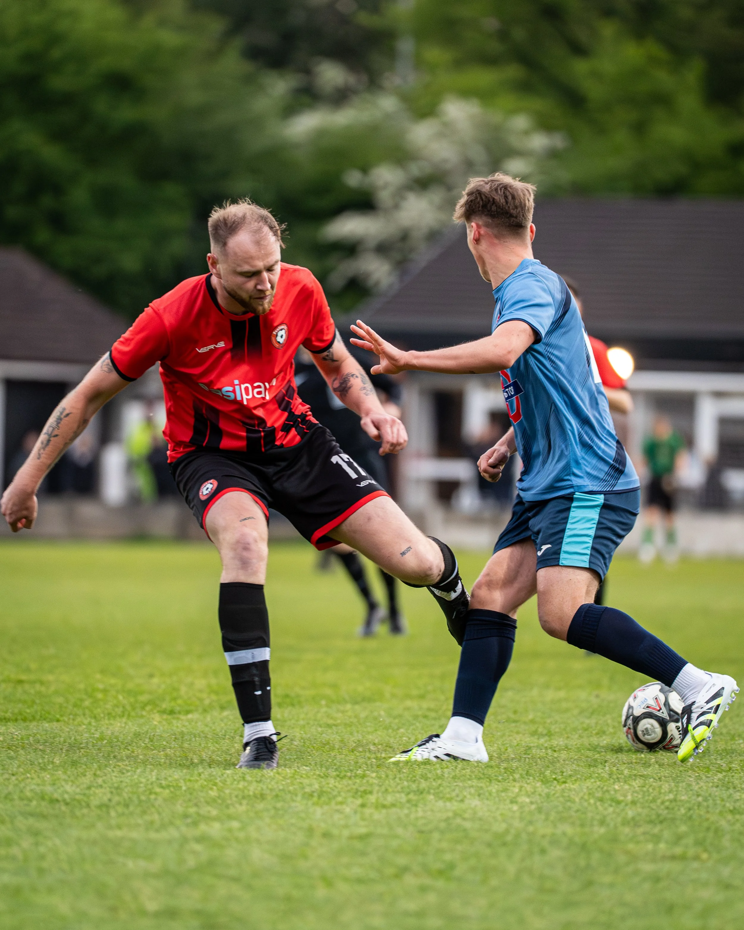 Two soccer players competing for the ball on a grassy field, one wearing a red and black jersey and the other in a blue jersey.