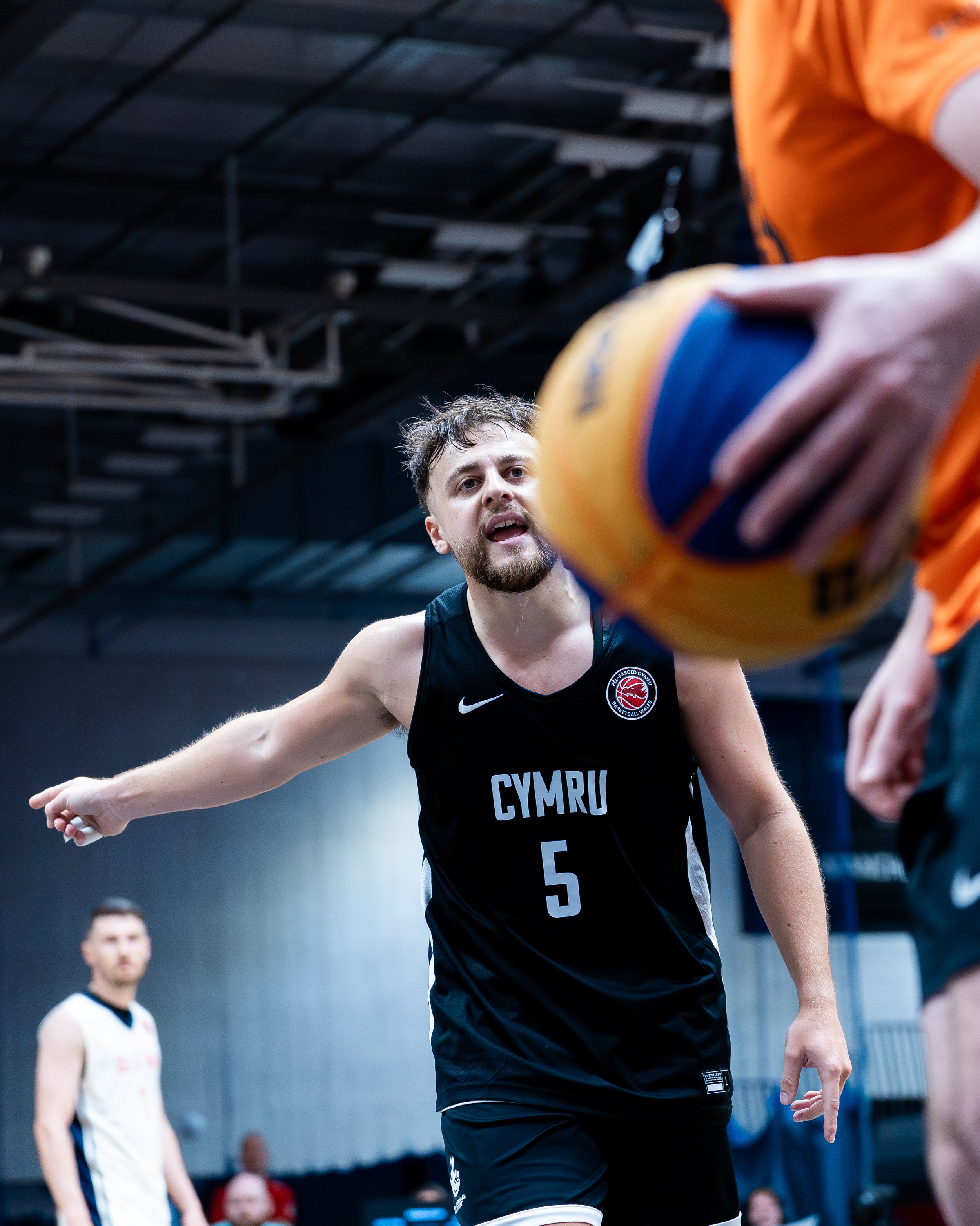 A male volleyball player with a beard, wearing a black jersey with 'CYMRU' and the number 5, is reaching out with his arm during a game in an indoor sports facility. Another player in a white jersey is in the background.