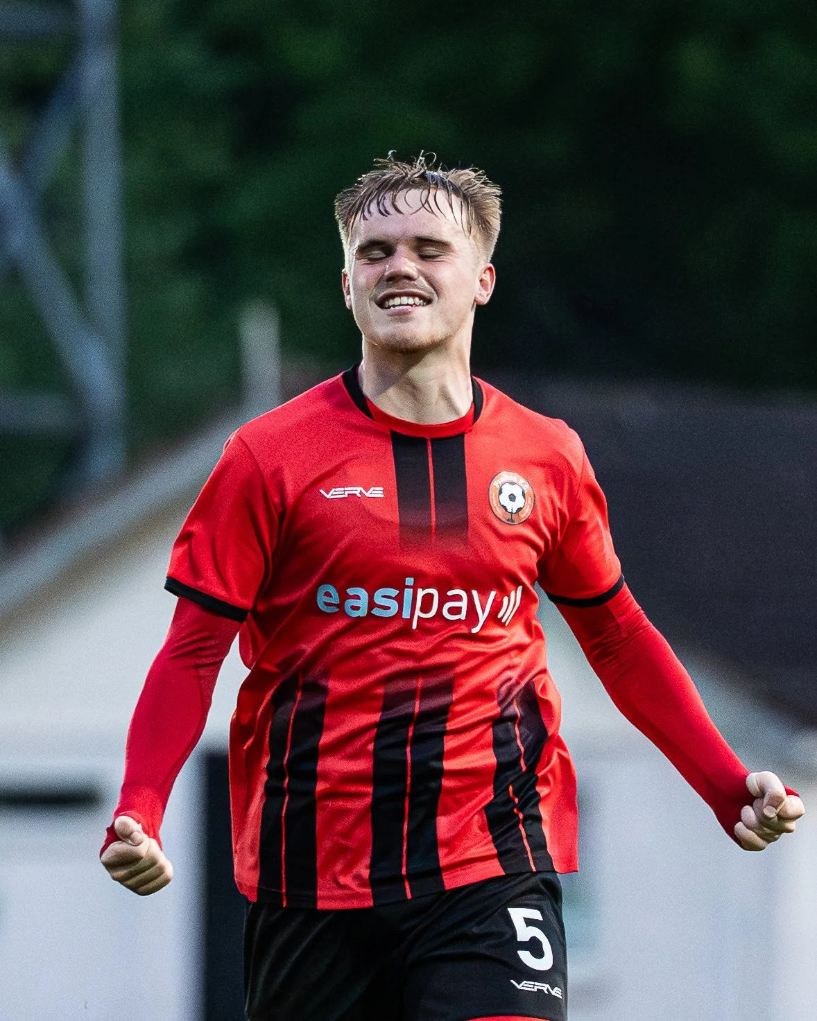 A young male soccer player with short blonde hair celebrating on the field, wearing a red and black jersey with the number 5 and various sponsor logos.