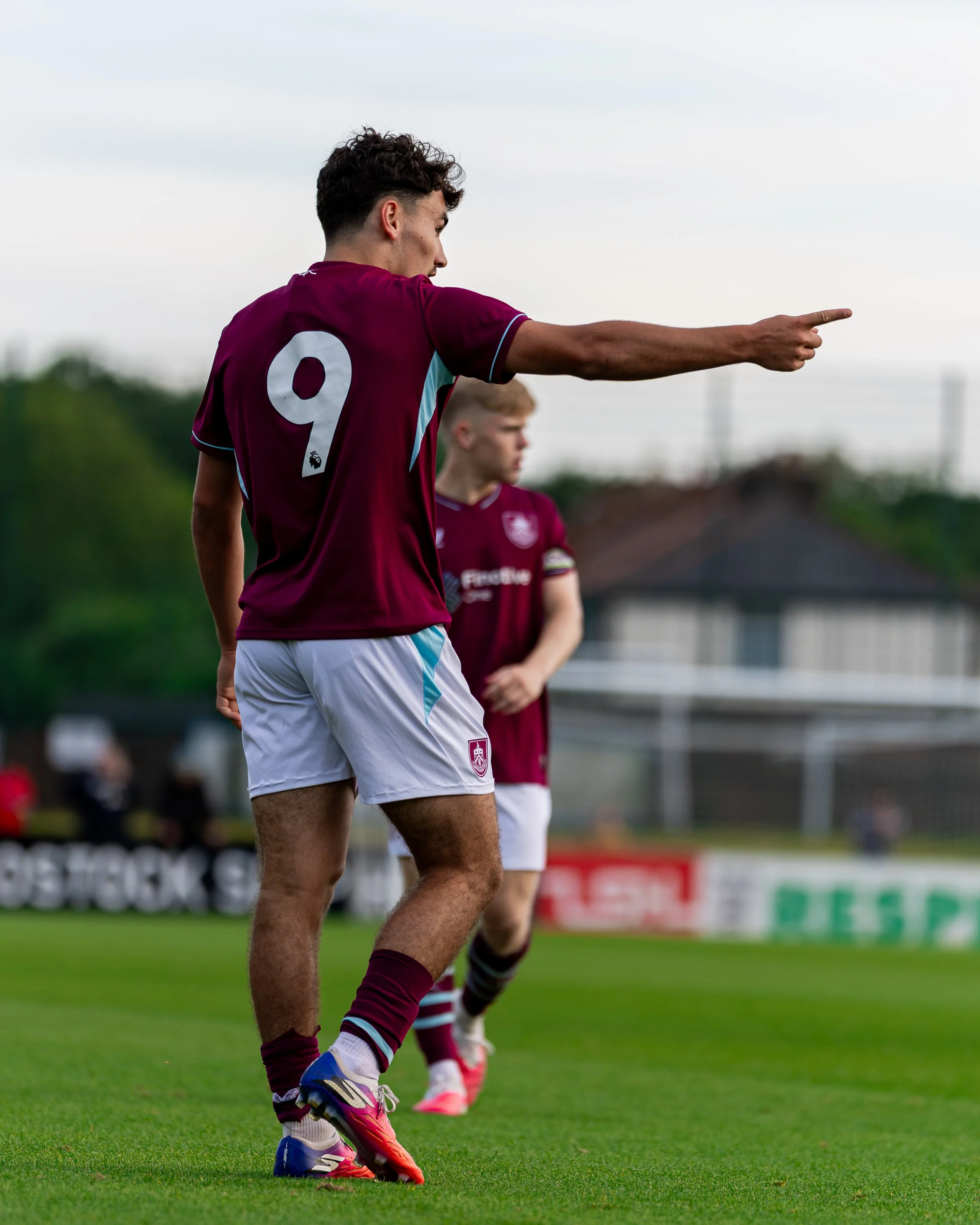 A soccer player wearing a maroon jersey with the number 9 points in a direction during a game on a grassy field, with another player in the background.