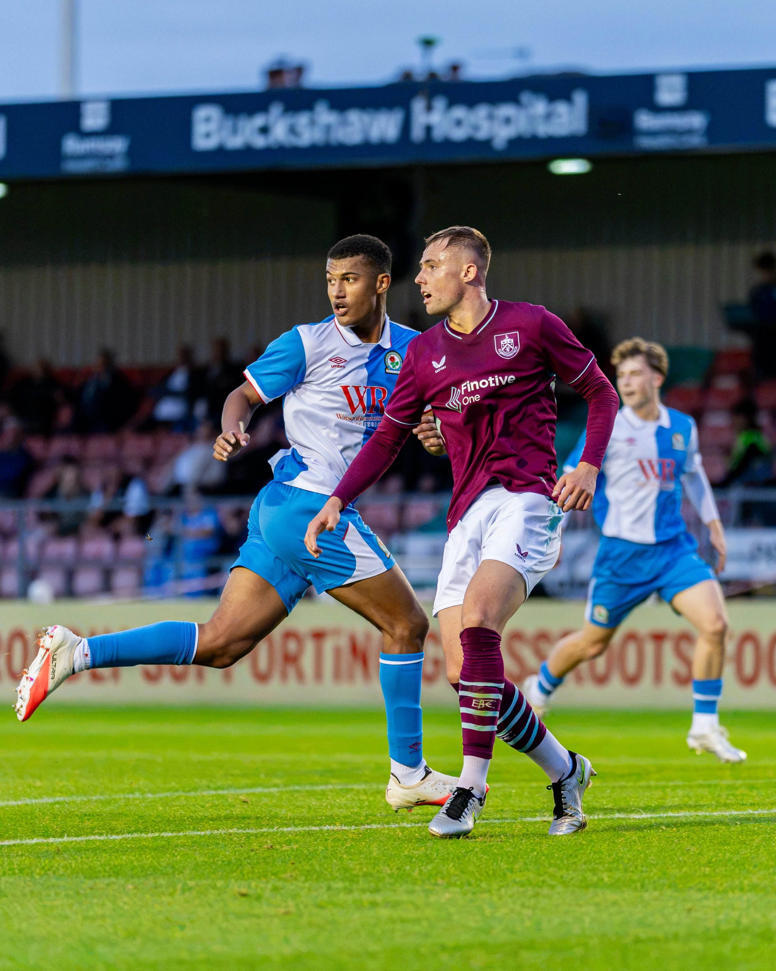 Two soccer players, one in a blue and white uniform and the other in a maroon uniform, competing for the ball on the field during a game at Buckshaw Hospital stadium.