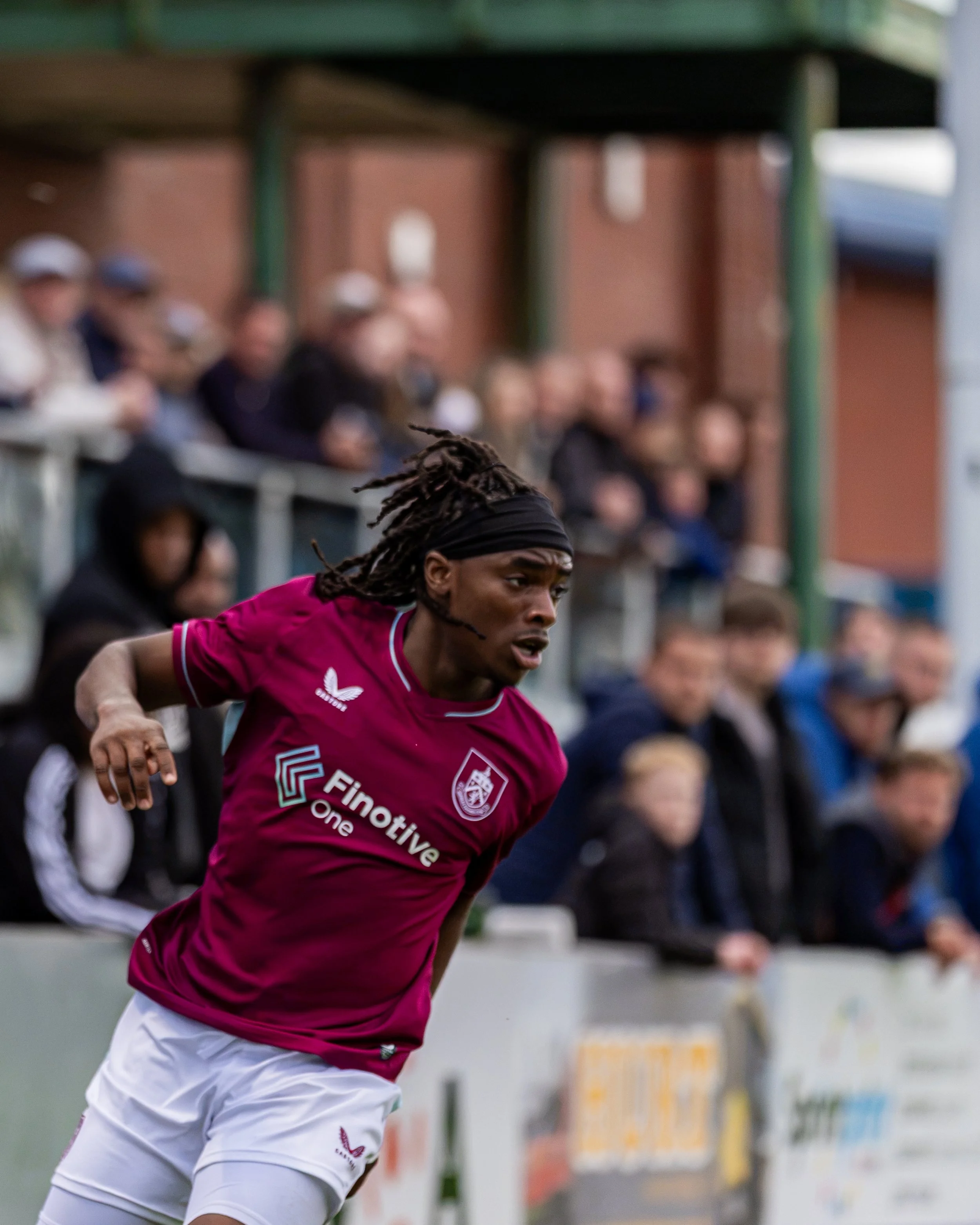 A man with dreadlocks wearing a pink sports jersey and white shorts, running on a sports field during a game, with spectators in the background.