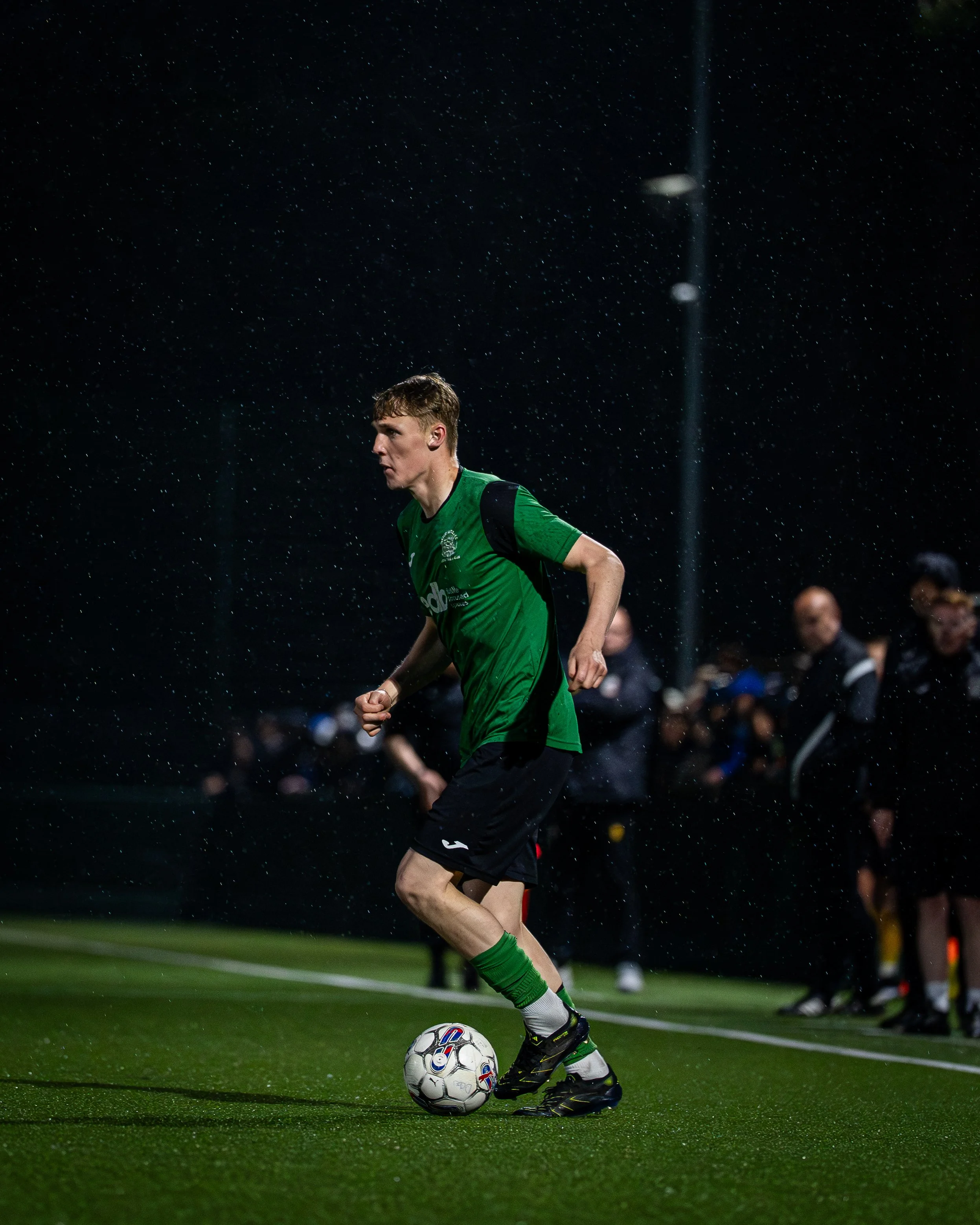A male soccer player in a green jersey and black shorts is about to kick the soccer ball on a rain-soaked field at night, with spectators and other players in the background.