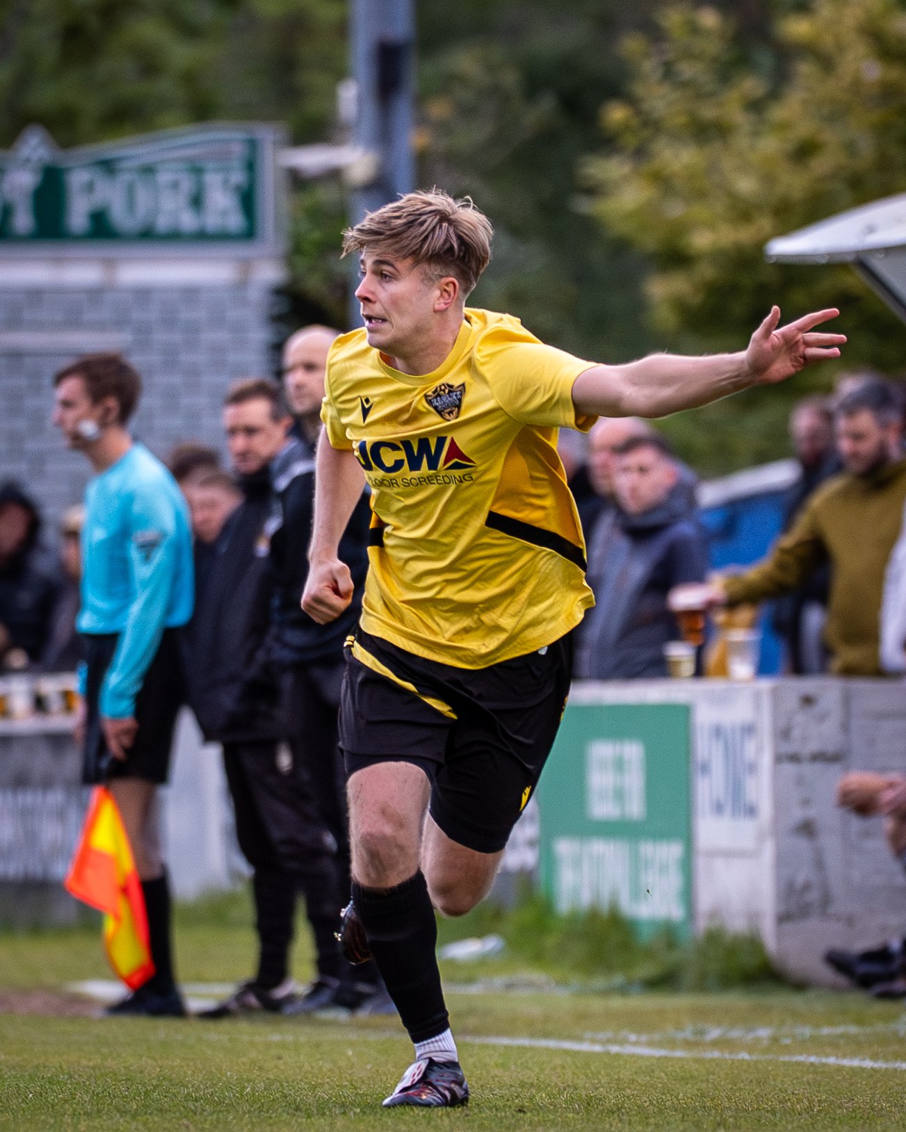 A young male soccer player in a yellow jersey and black shorts is celebrating on the field with outstretched arms, during a match. Spectators are seen in the background.