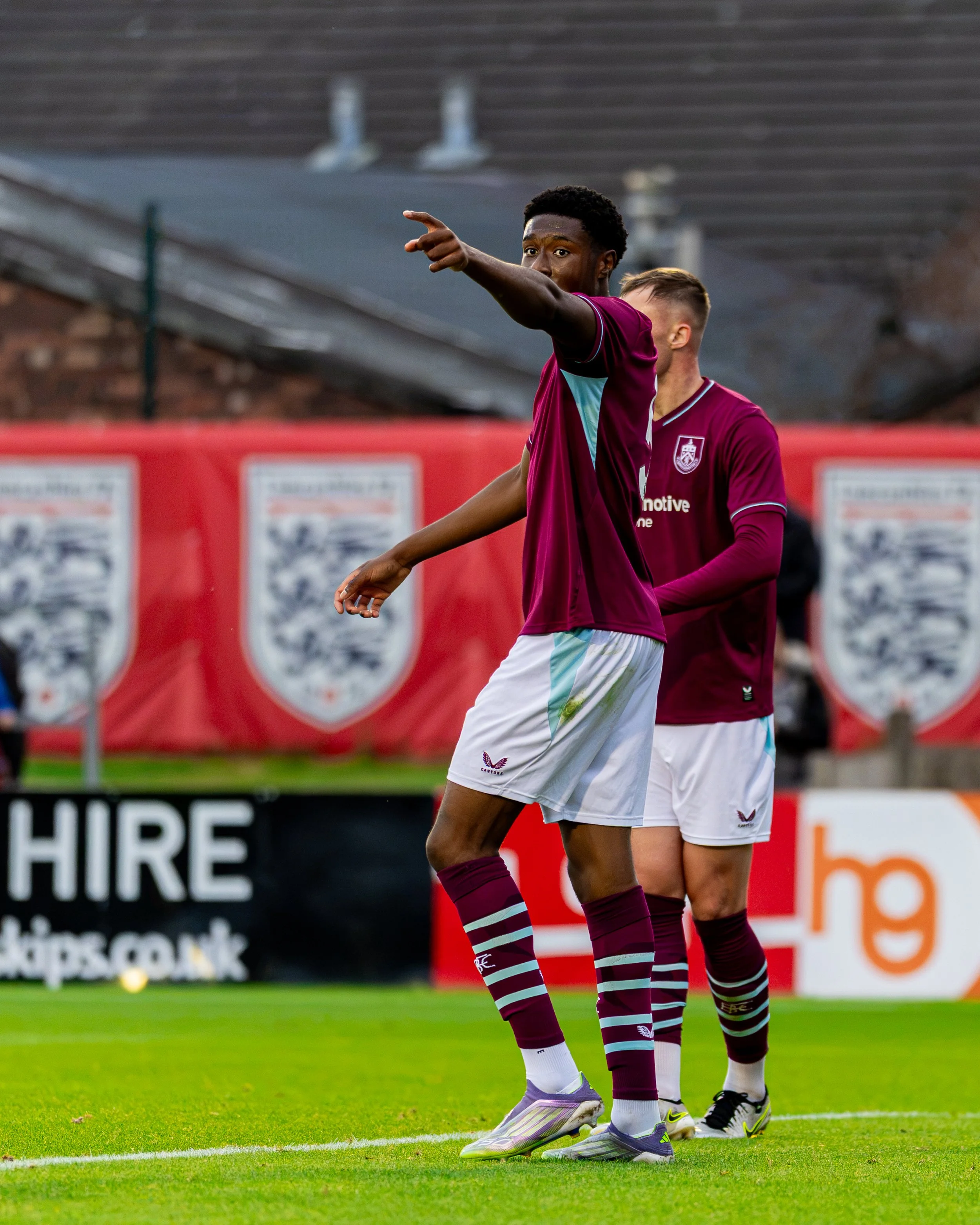 Soccer players on the field, one pointing forward, wearing maroon and blue uniforms with striped socks.
