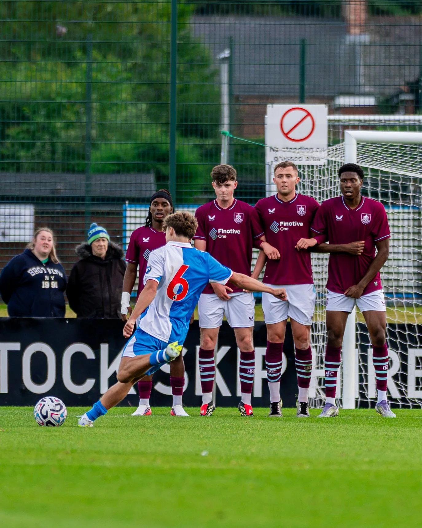 A soccer player wearing a blue and white uniform with the number 6 on his back is kicking a soccer ball. Behind him, four players in maroon uniforms are standing near the goal, with some observing the game. A few spectators are visible on the sidelin