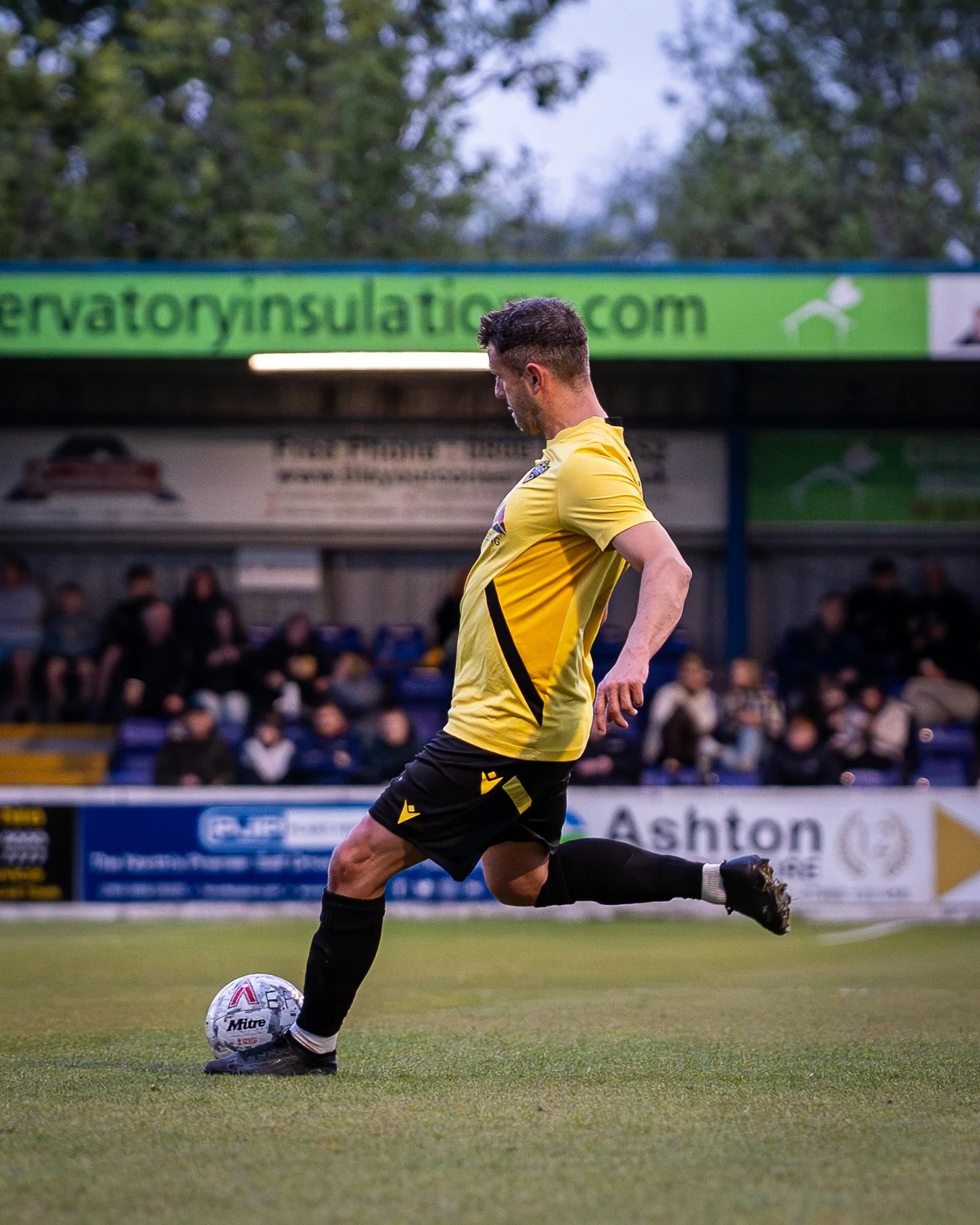 A soccer player in yellow jersey and black shorts kicking a white soccer ball on a field during a game with spectators in the background.