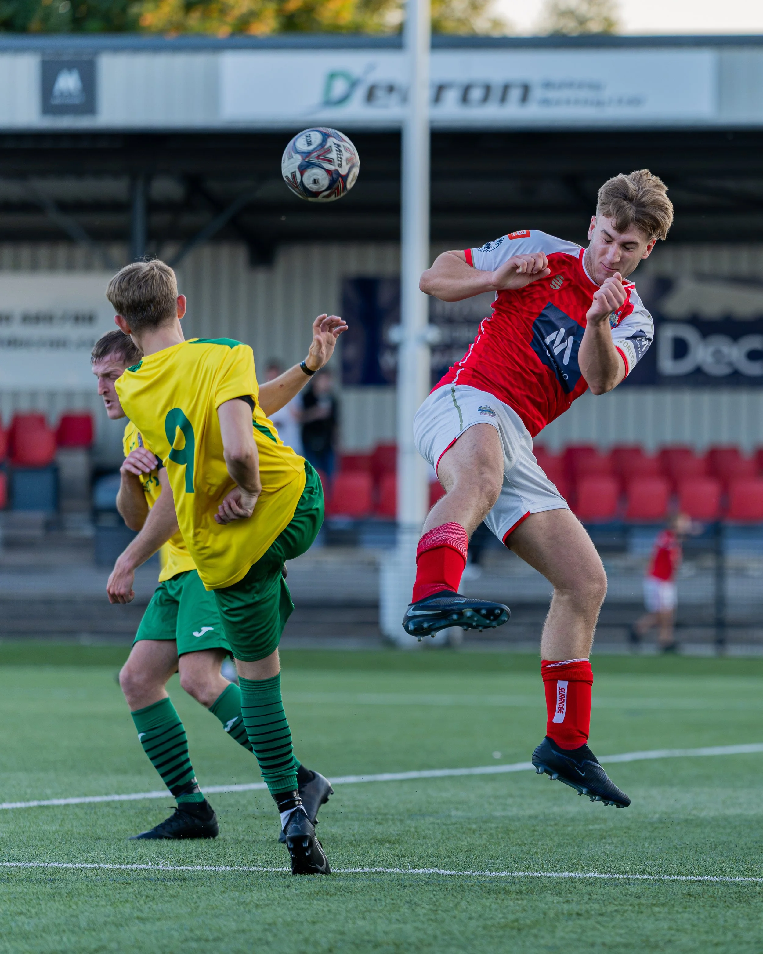 Young male soccer players competing for the ball during a match on a green field, with one player in a red and white uniform and another in a yellow and green uniform, under clear daylight.