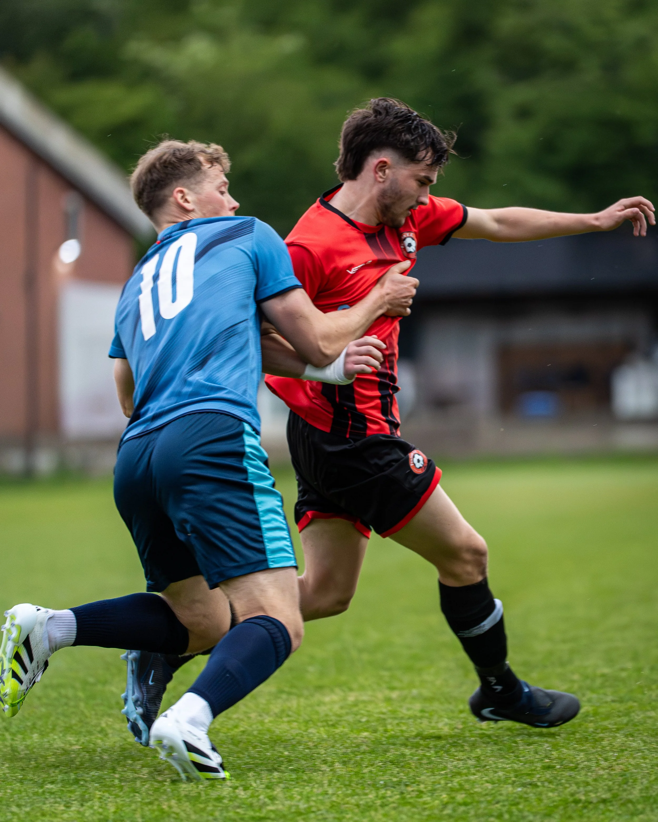 Two male soccer players in action during a match, running on a green grass field with trees in the background.