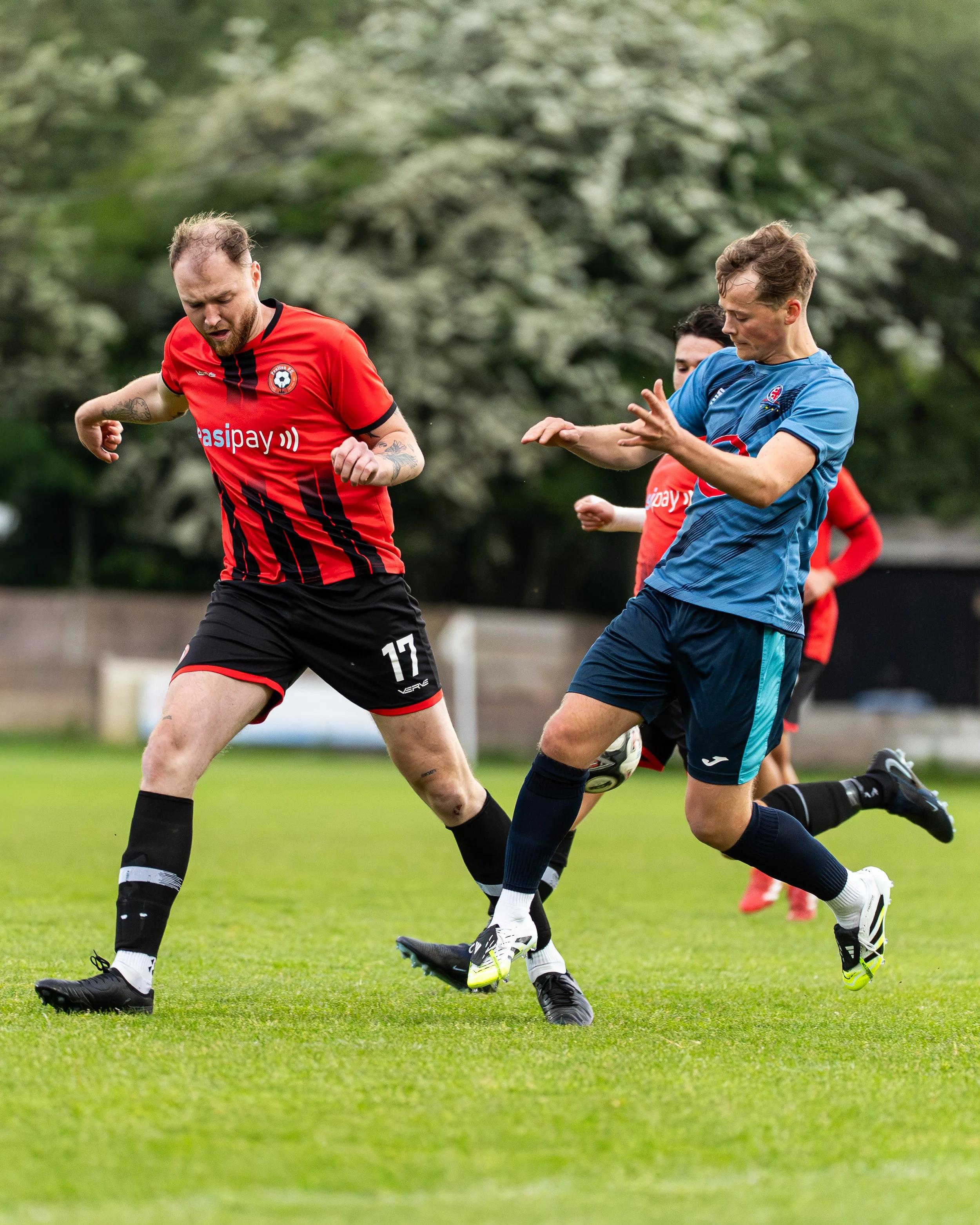 Two male soccer players are competing for the ball on a grassy field, with a blurred background of trees. One player is wearing a red and black kit, and the other is in a blue kit. They are both in motion, focusing on the ball, with their legs and ar