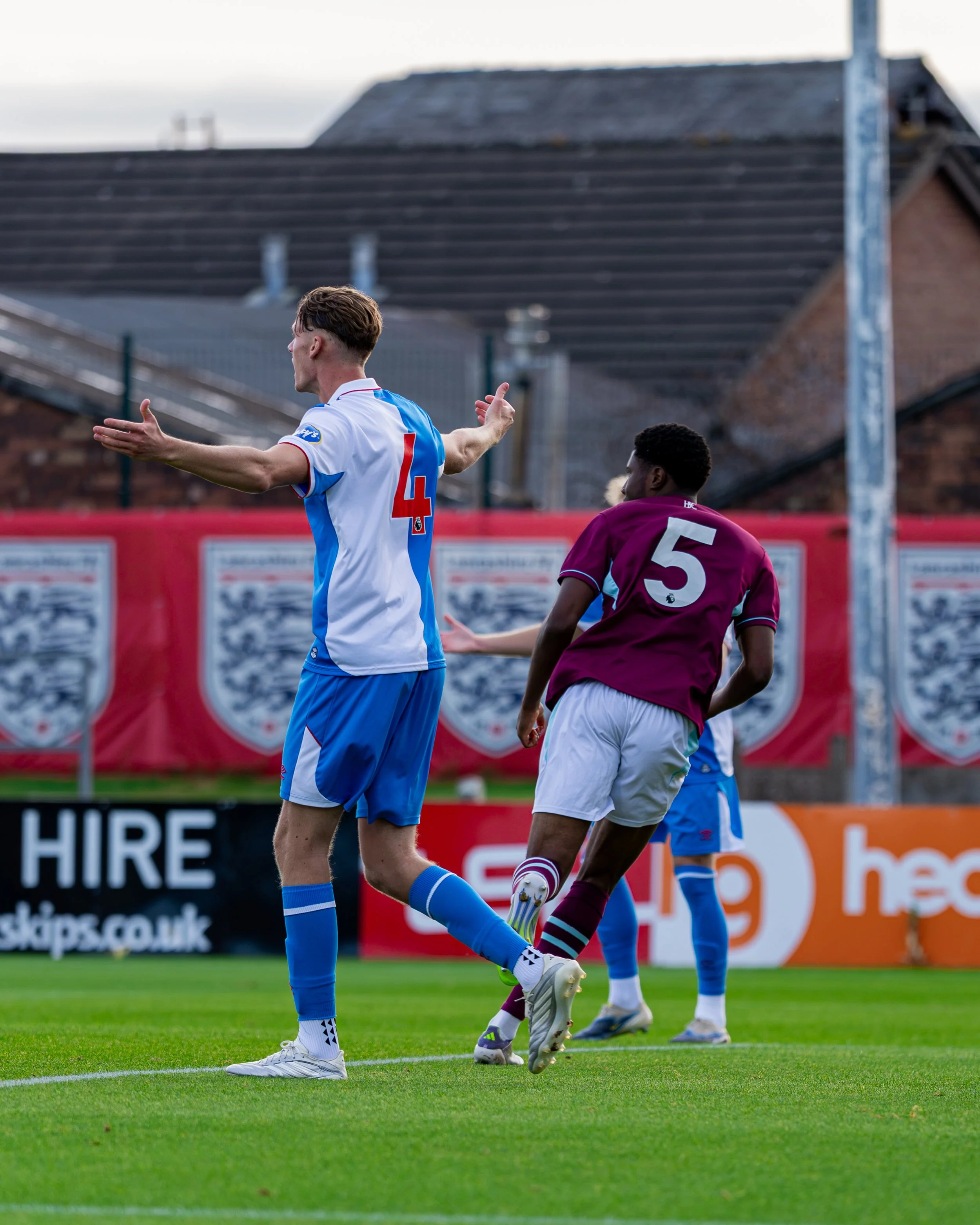 Two soccer players are on the field during a game, with one celebrating with open arms and the other looking at the ball. The player in the foreground wears a white and blue jersey with the number 4, while the player in the background wears maroon wi