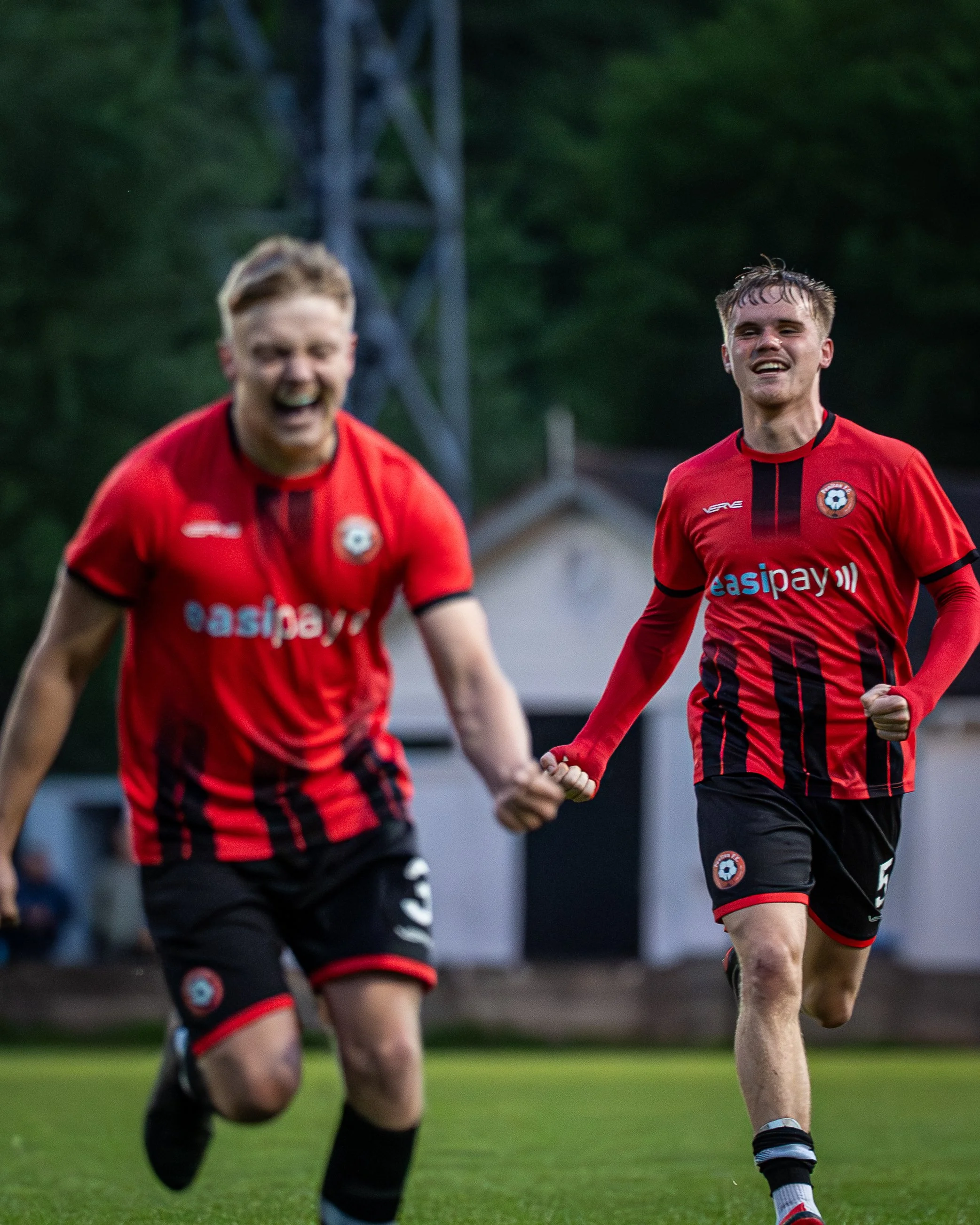 Two soccer players in red and black uniforms celebrating on the field, one smiling and holding hands, the other laughing with mouth open, green trees and a structure in the background.