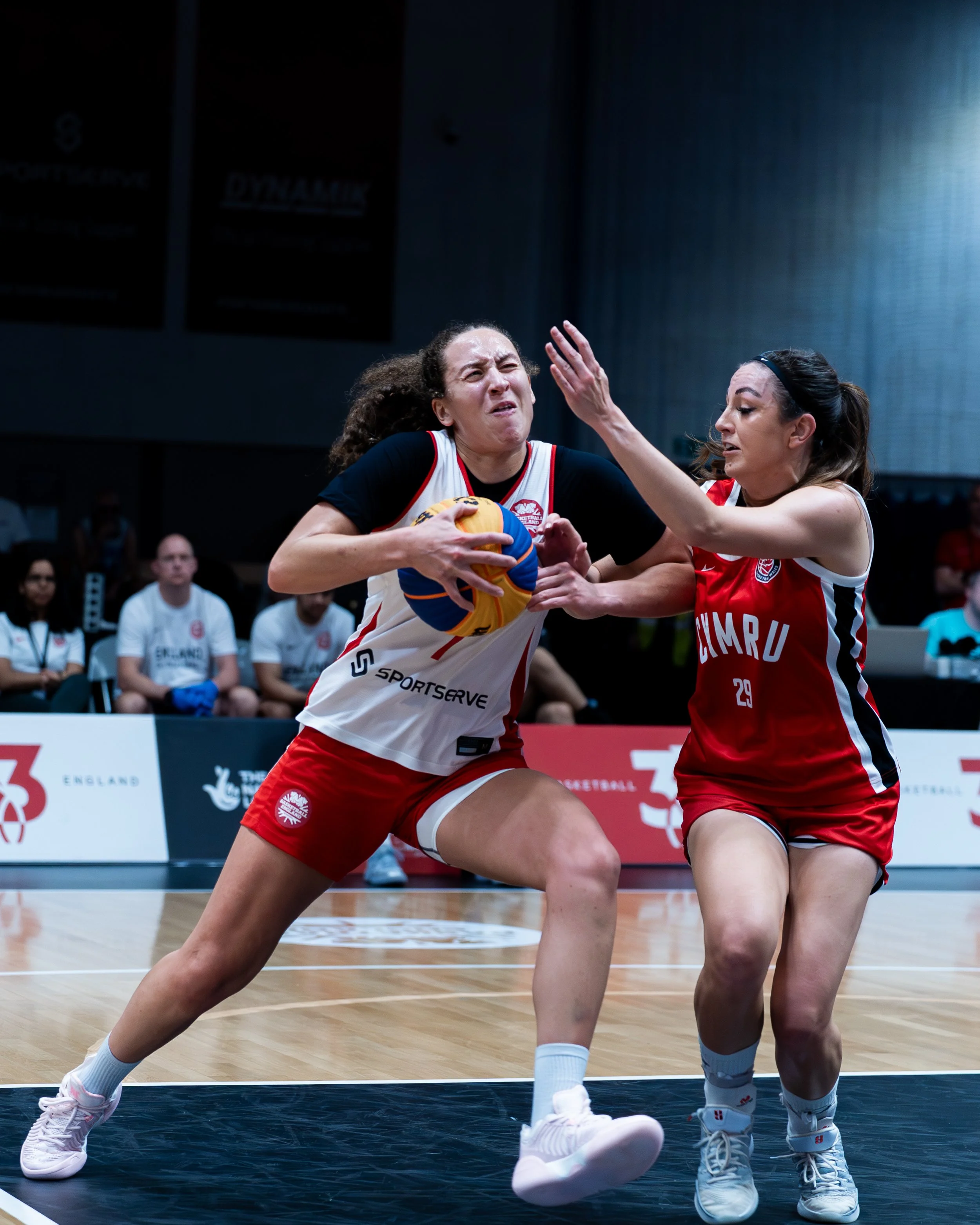 Two female basketball players fighting for possession of a basketball during a game, with a basketball court and audience in the background.