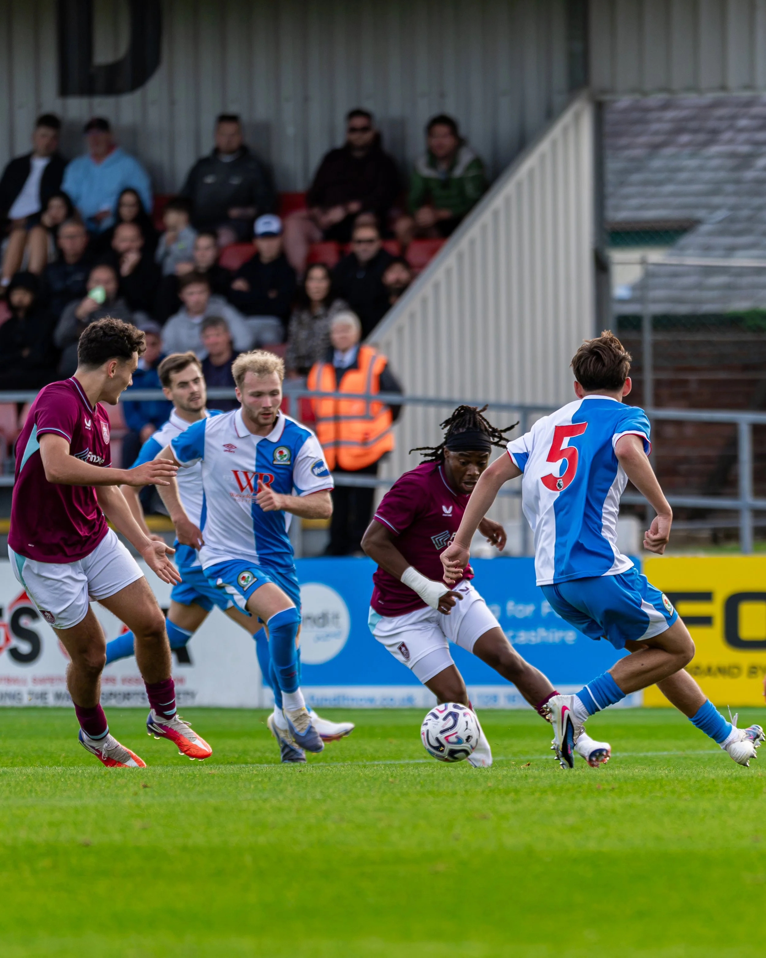 Soccer players in maroon and blue jerseys compete for ball on field with audience in background.