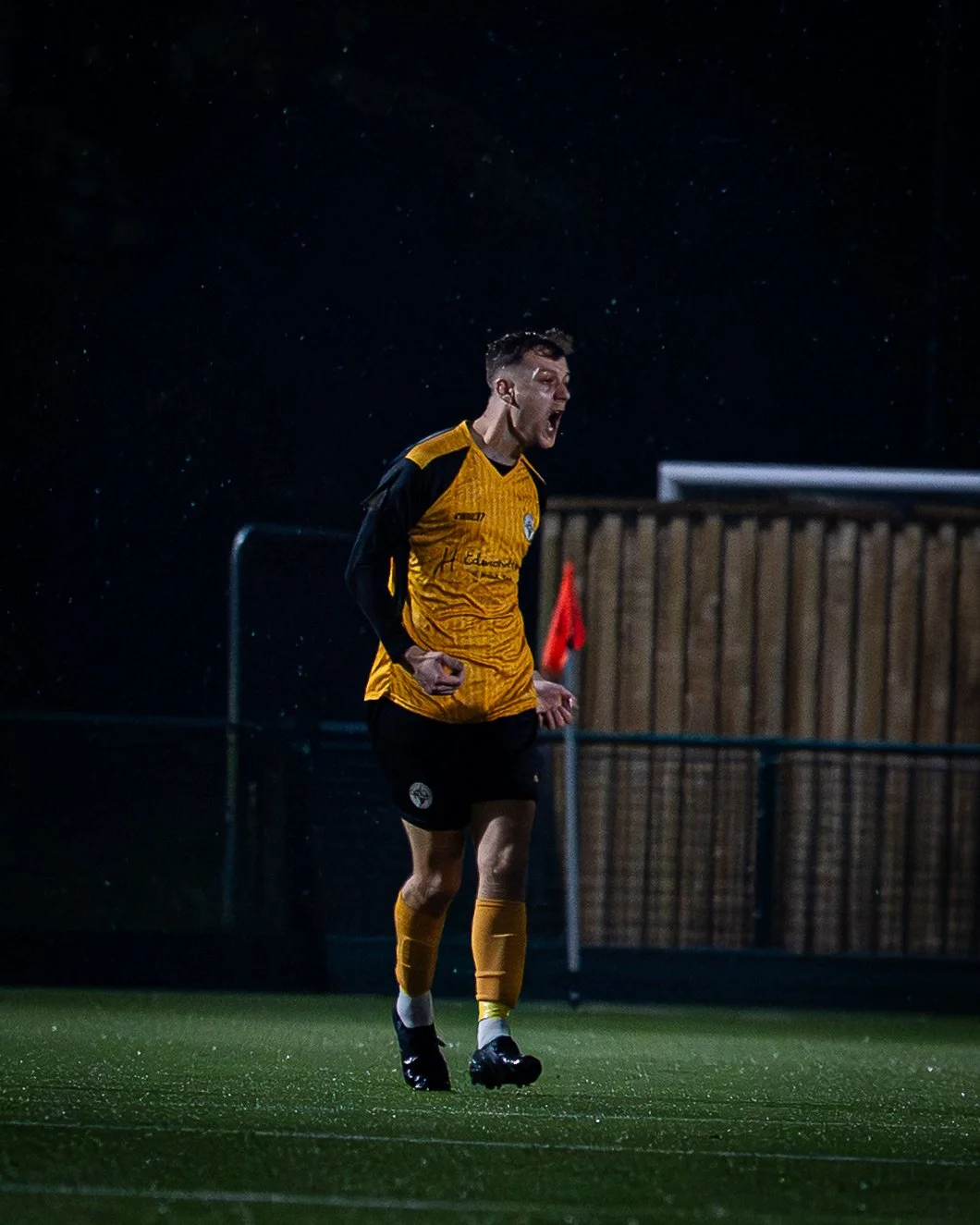 A soccer player wearing a yellow and black jersey is celebrating on the field at night, with rain falling.