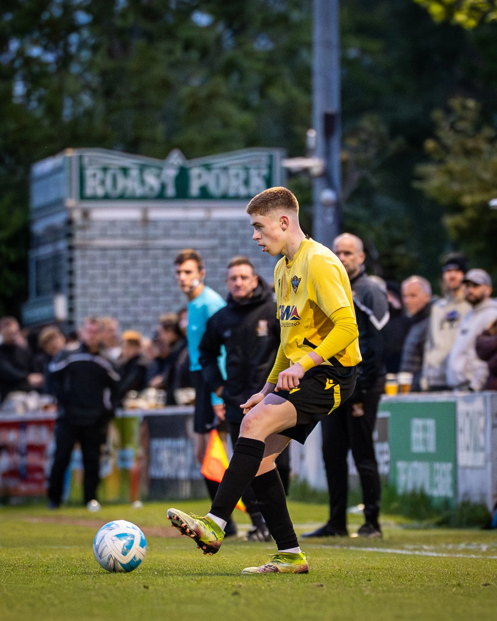 A young male soccer player wearing a yellow jersey, black shorts, and neon green cleats kicks a white and blue soccer ball during a game. Several spectators are visible in the background, watching the match on a grass field with trees and a sign that