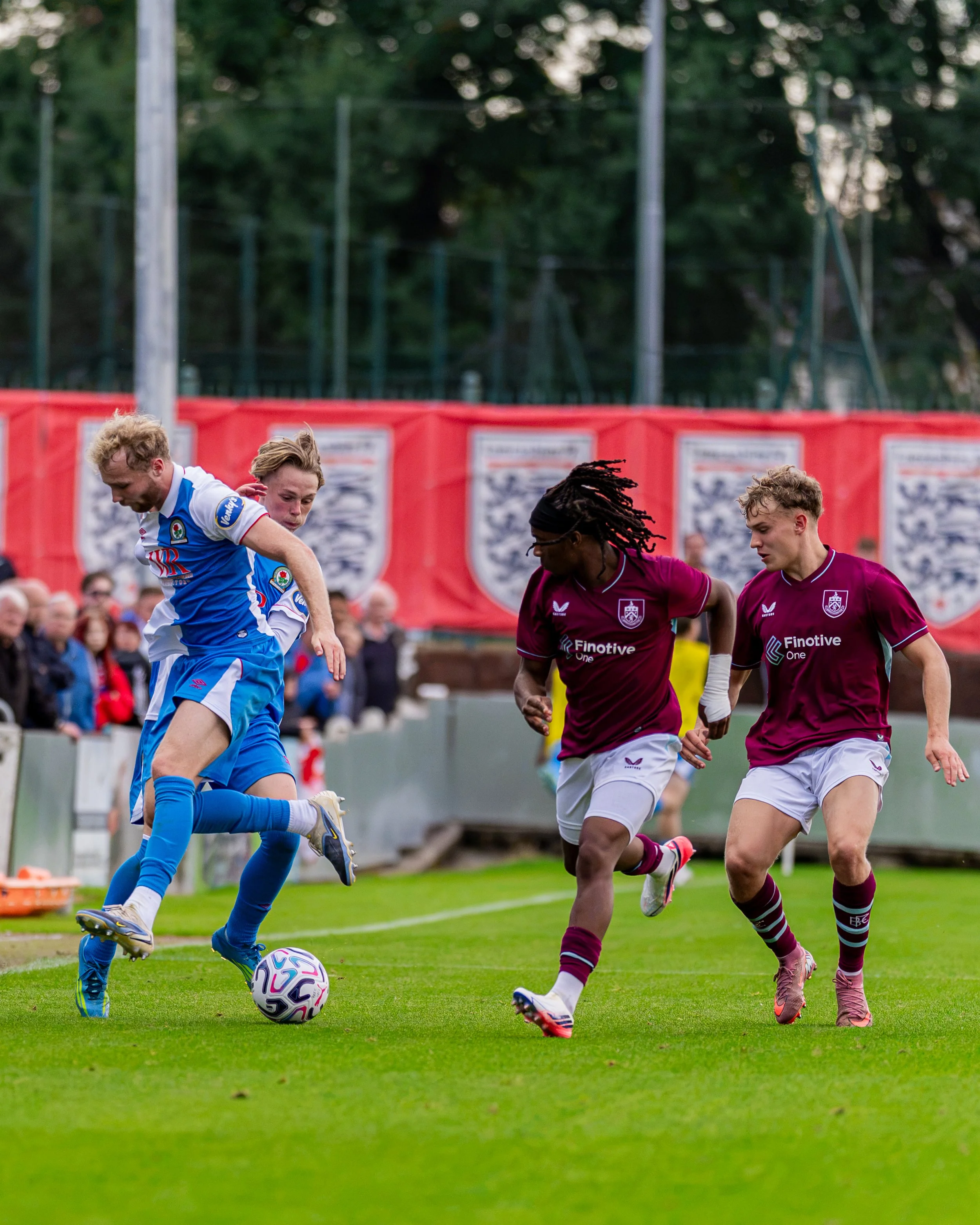 Four soccer players in action on a grass field, two wearing maroon jerseys and two wearing blue jerseys, with the player in the blue jersey about to kick the ball, and an audience in the background.