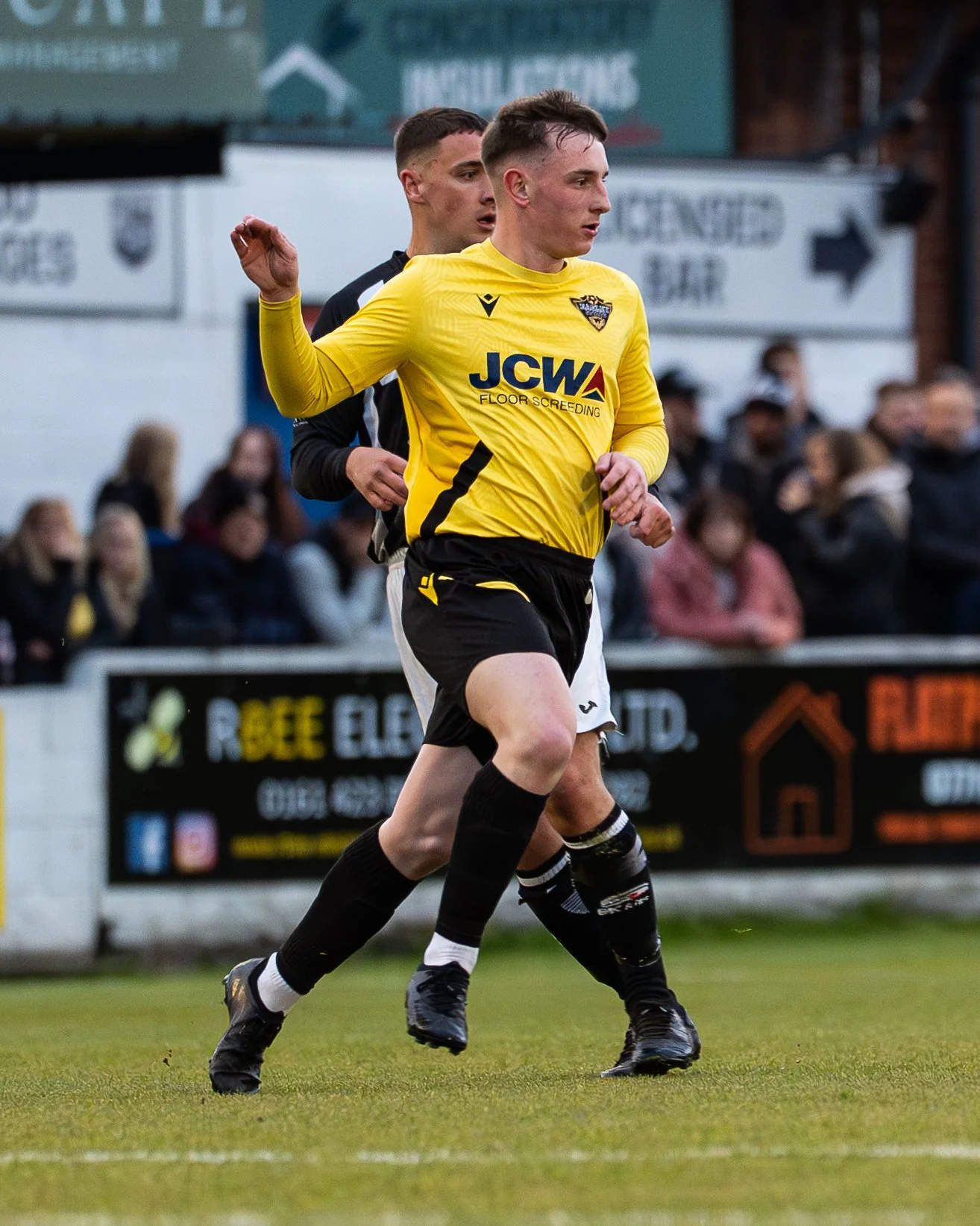 Two soccer players running on the field during a game, one wearing a yellow jersey and the other in black, with spectators in the background.