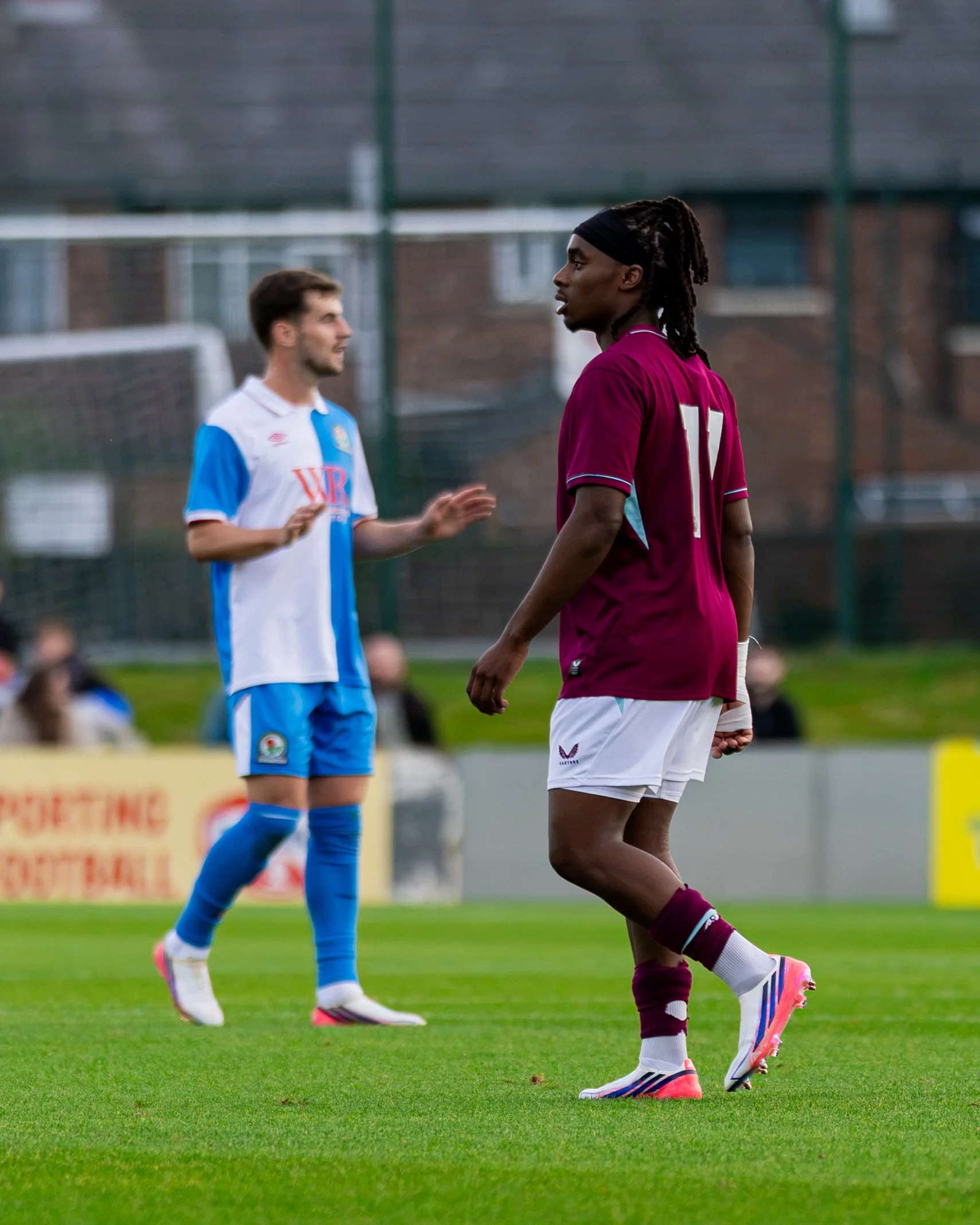 Two male soccer players on a green field during a match, one in a maroon jersey and white shorts, and the other in a blue and white jersey with shorts, engaging in play.
