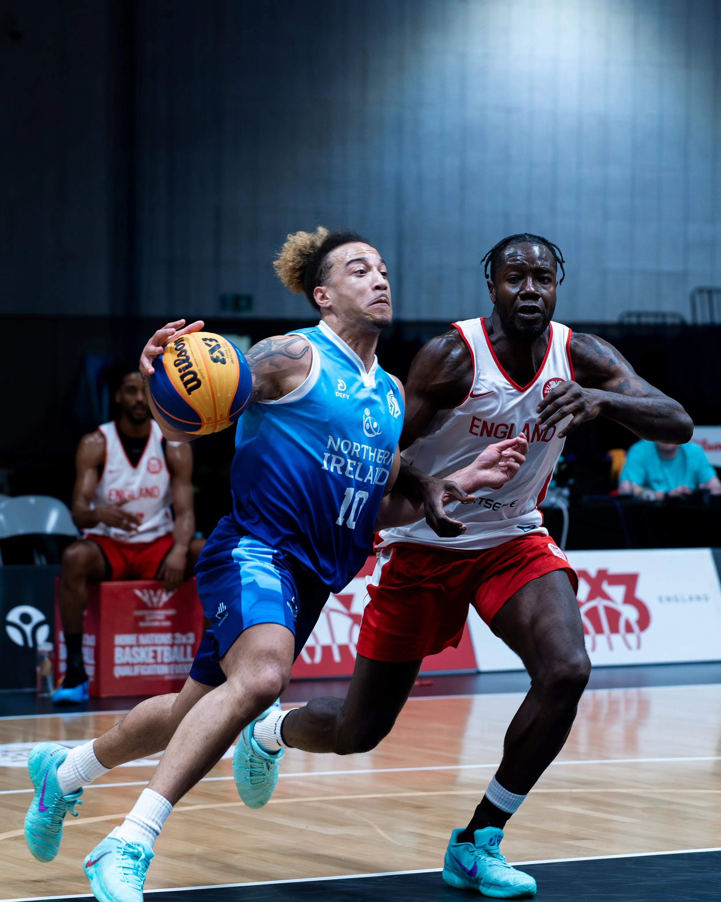 Two basketball players from opposing teams competing for the ball on a basketball court. The player on the left, wearing a blue jersey with the text 'Northern Ireland,' is holding the ball, while the player on the right, in a white and red jersey wit
