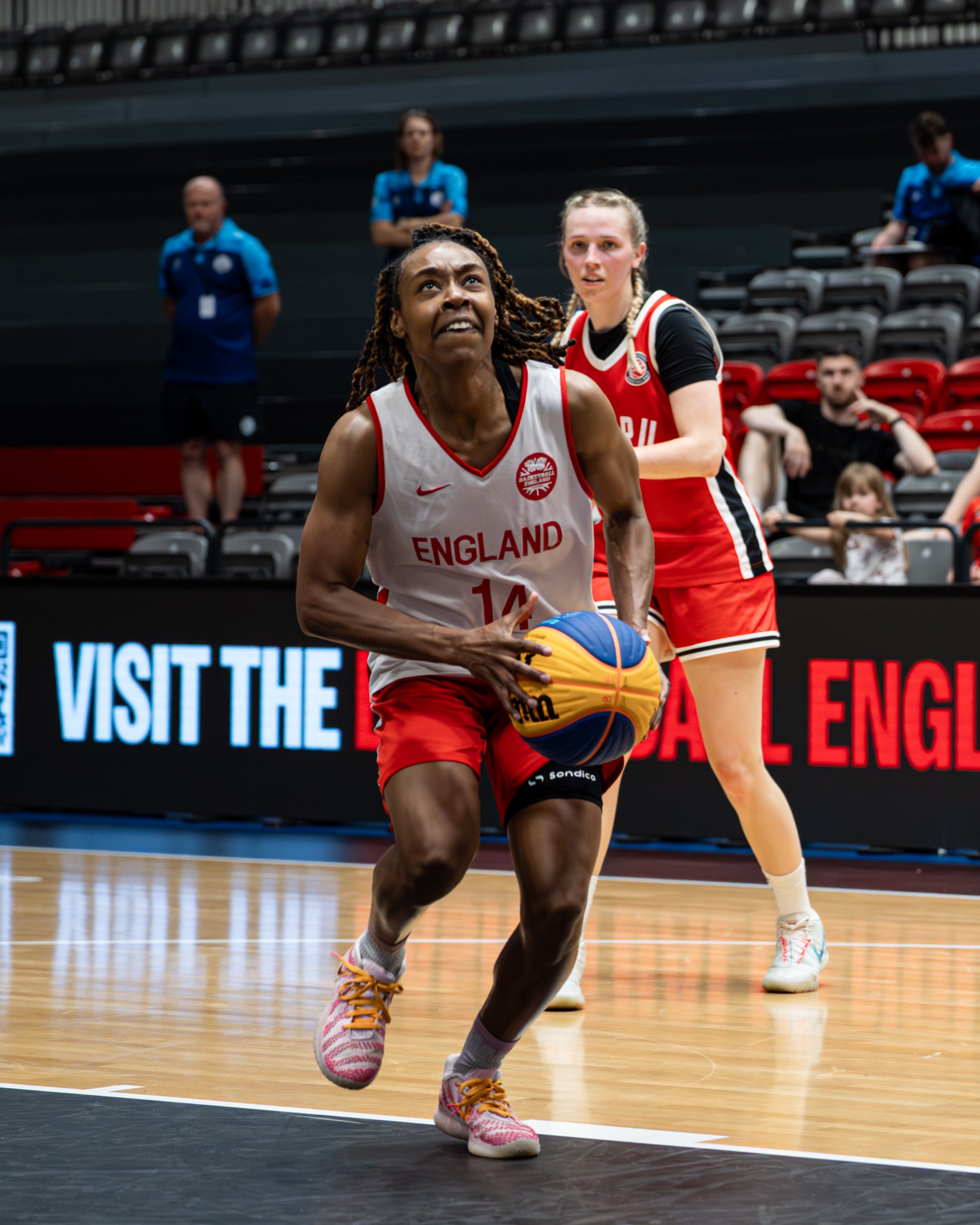 A female basketball player from England with dreadlocks, wearing a white jersey with red trim, dribbling a basketball on an indoor court. Another female player in a red jersey is in the background, and spectators and officials are visible.