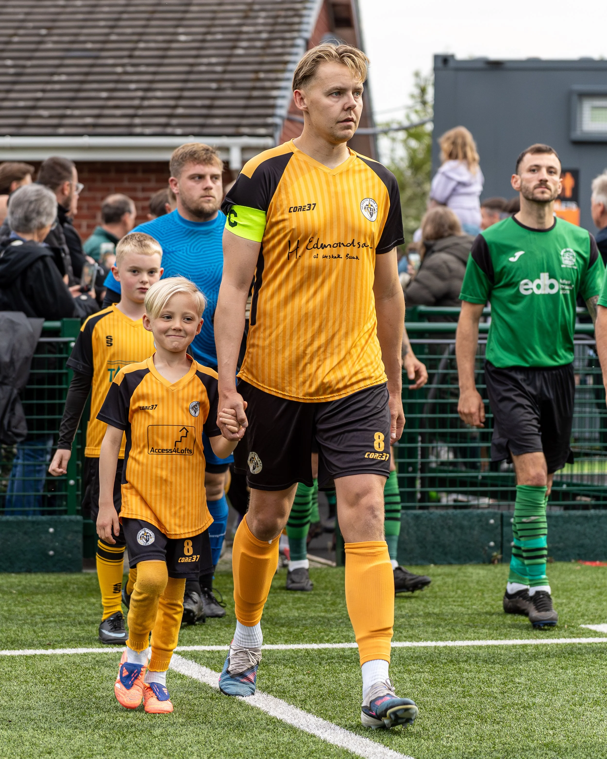 Soccer players, including a tall man in a yellow and black uniform with the number 8, walking onto the field with children, spectators, and other players in the background.