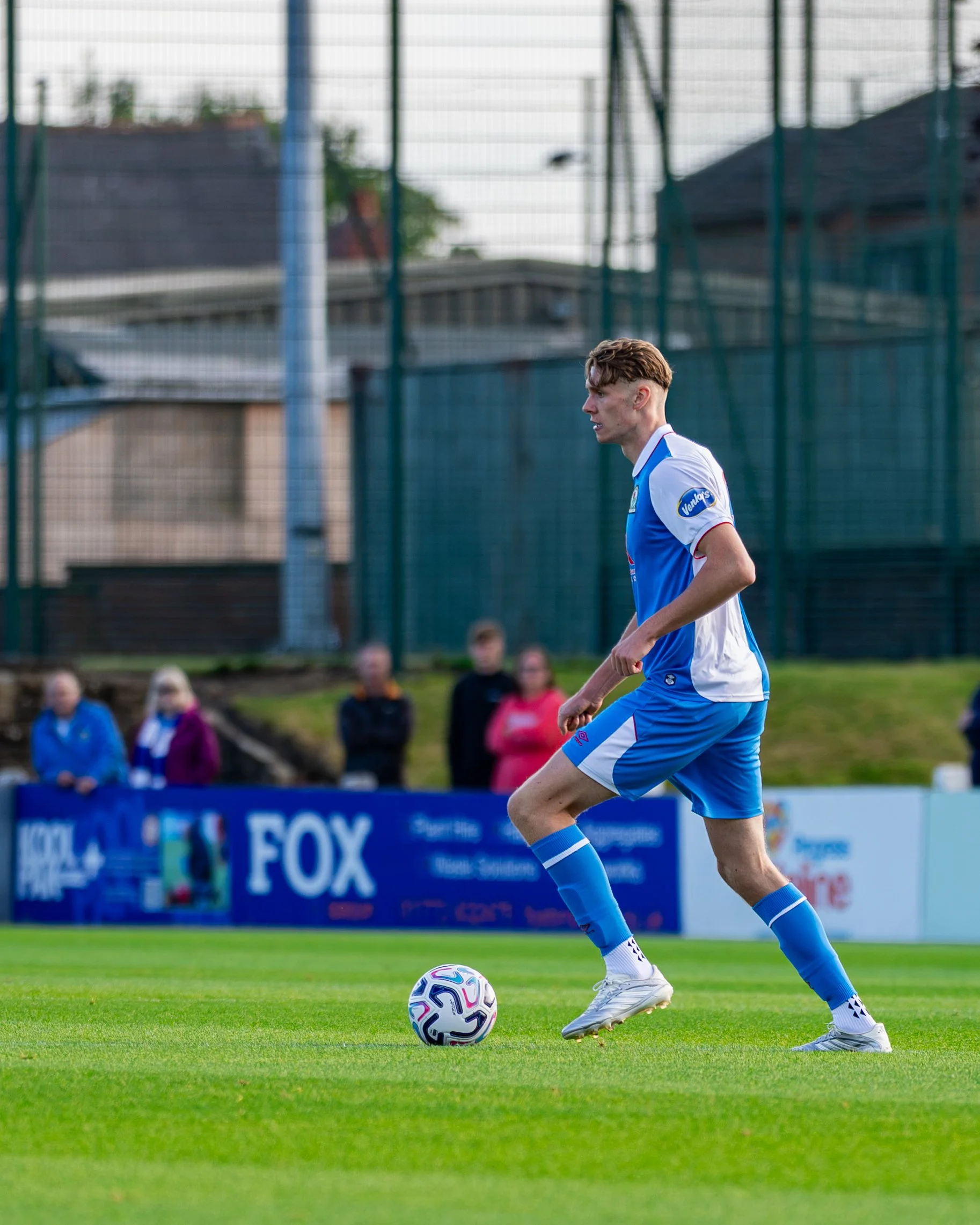 A male soccer player in a blue and white uniform is preparing to kick a soccer ball on a grassy field. In the background, there are spectators and a fence around the field.