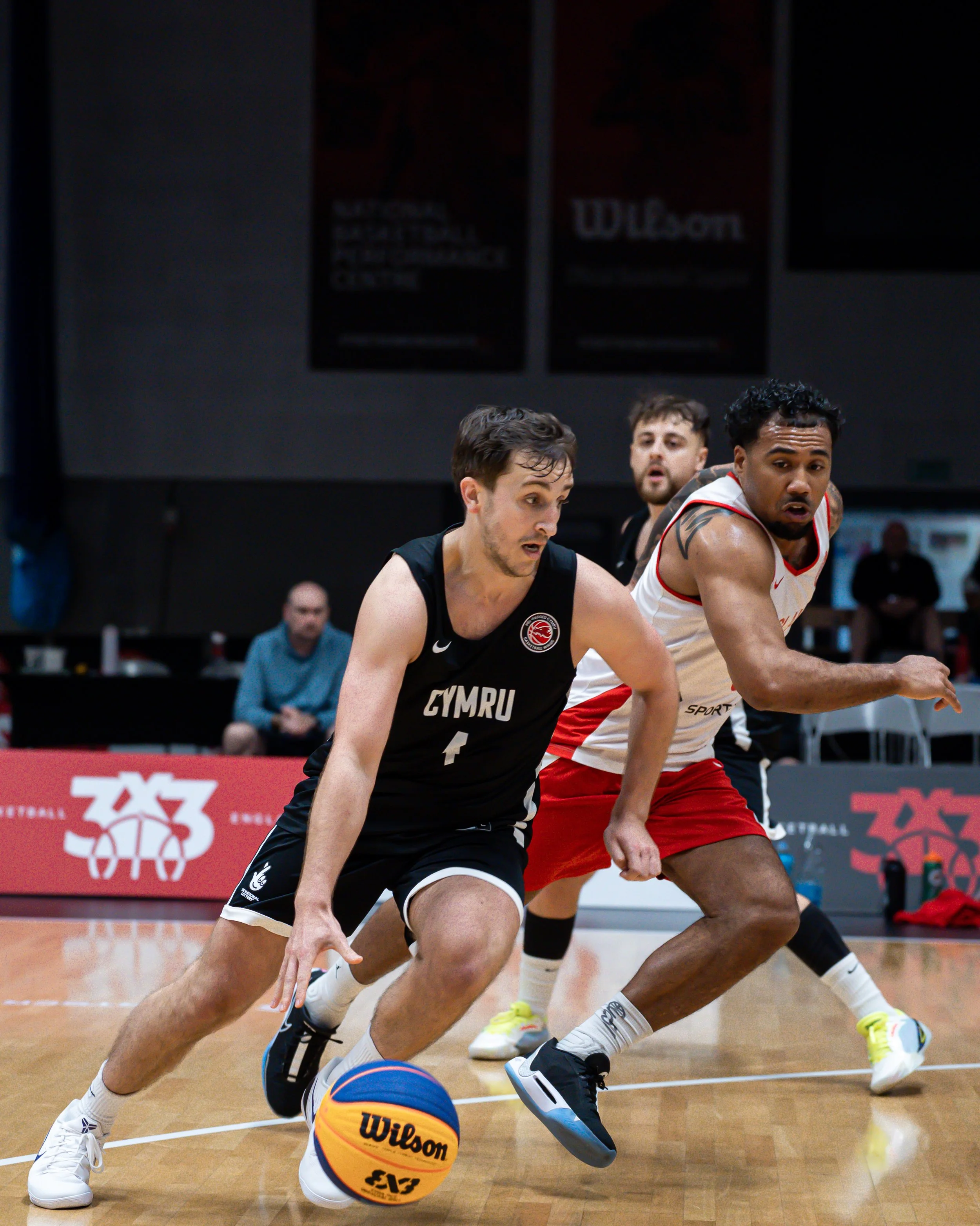 Two basketball players competing for the ball during a game on an indoor court, with spectators and banners in the background.