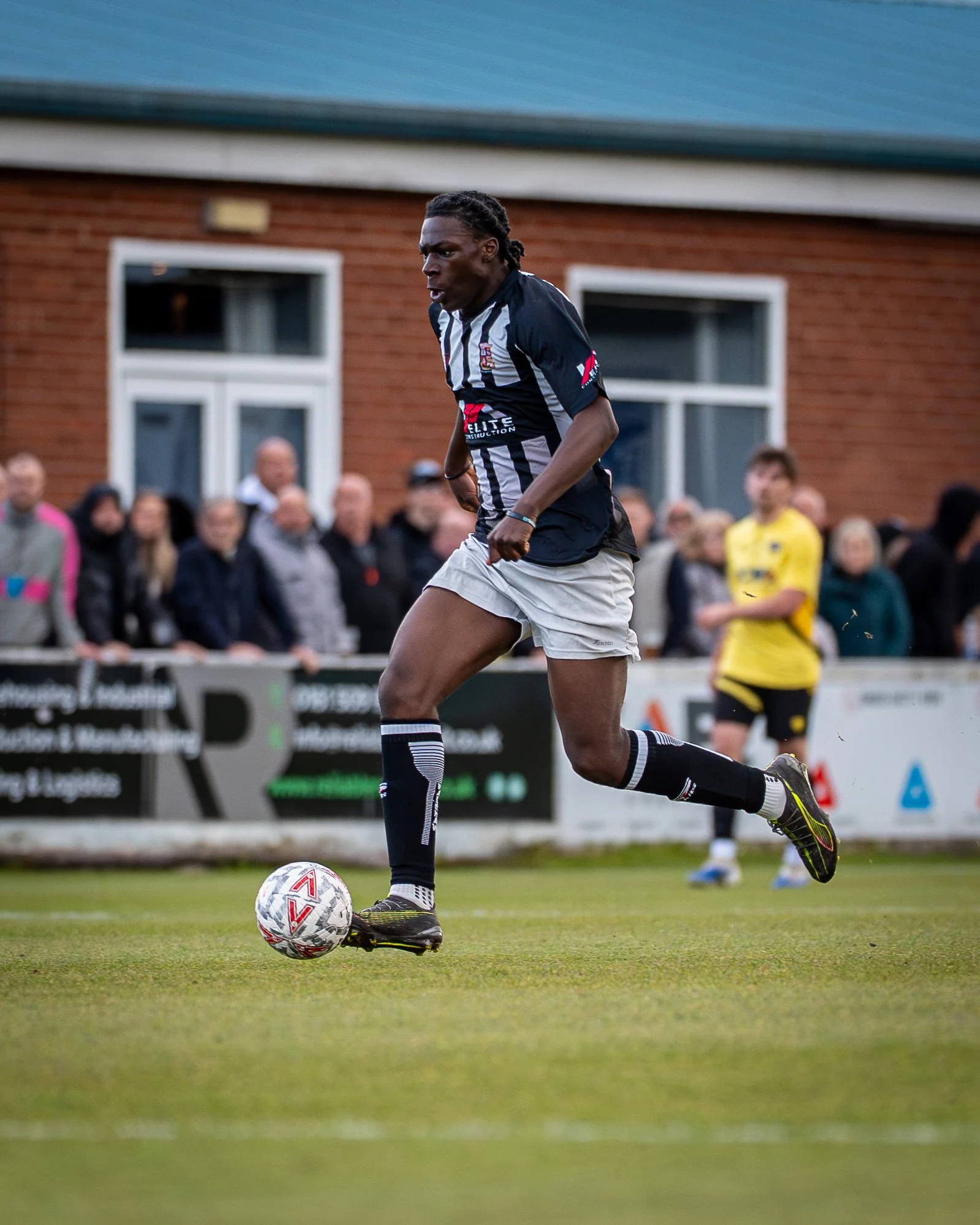 A football player wearing a black and white striped jersey, white shorts, and black socks is kicking a soccer ball on a grass field, with spectators and a building in the background.