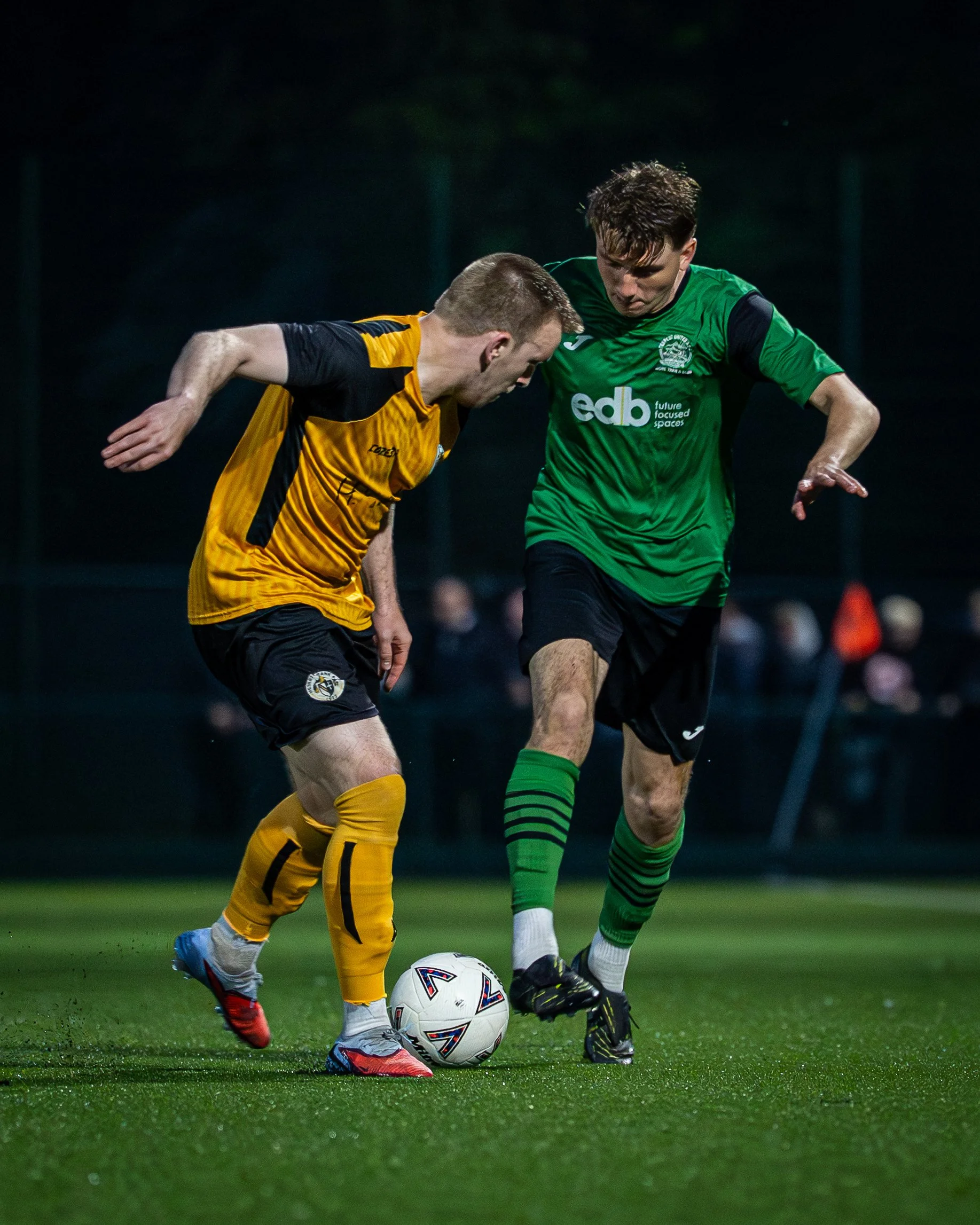 Two soccer players from opposing teams compete for the ball during a match at night. The player in yellow and black is kicking the ball, while the player in green is trying to intercept. Both players are focused on the ball, with a blurred background